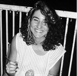 Mulher sorridente com cabelo cacheado, segurando uma taça, em uma área com grades ao fundo
