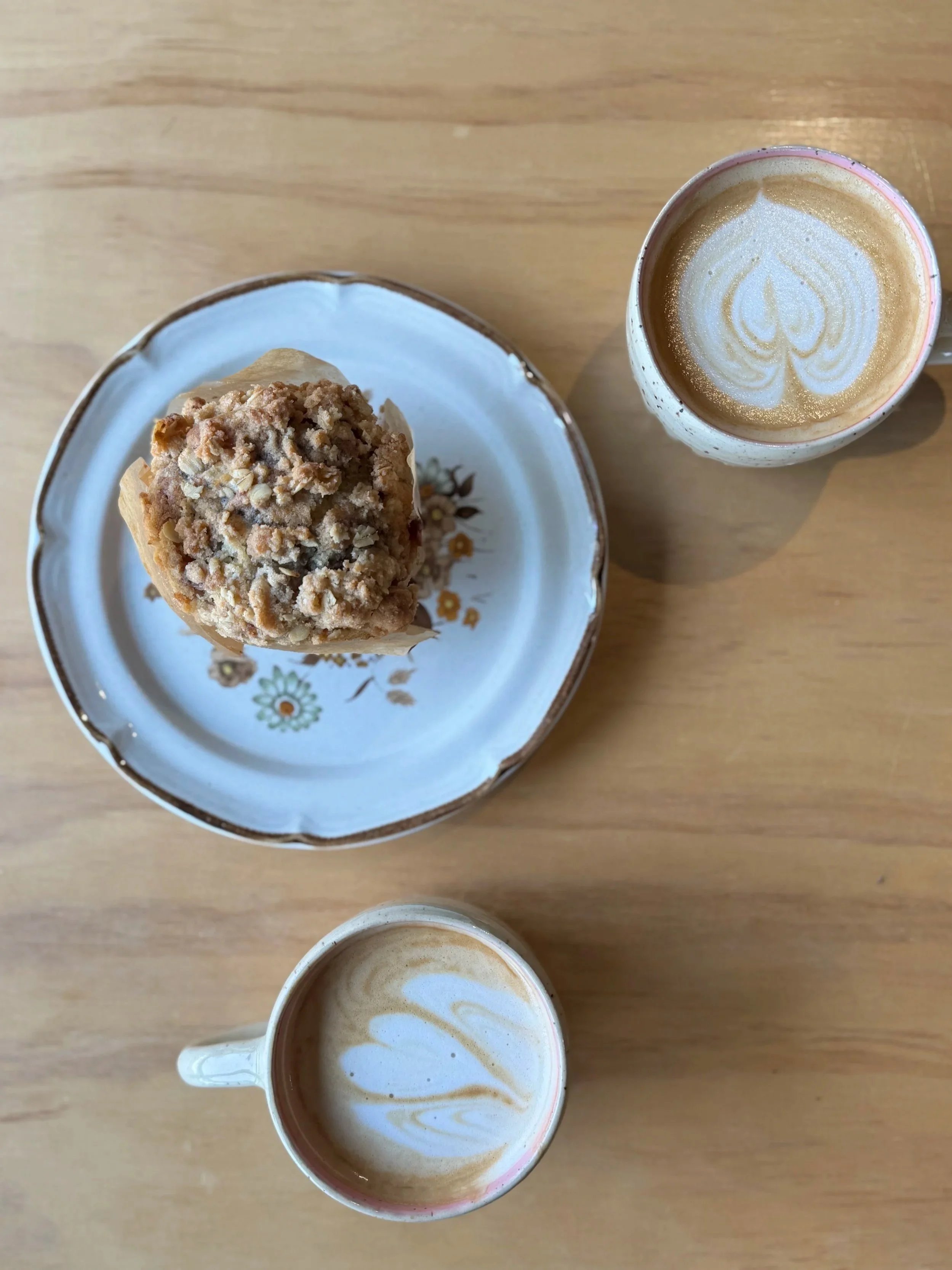 Top view of a muffin on a decorative plate, two cups of latte with latte art, all on a wooden table.