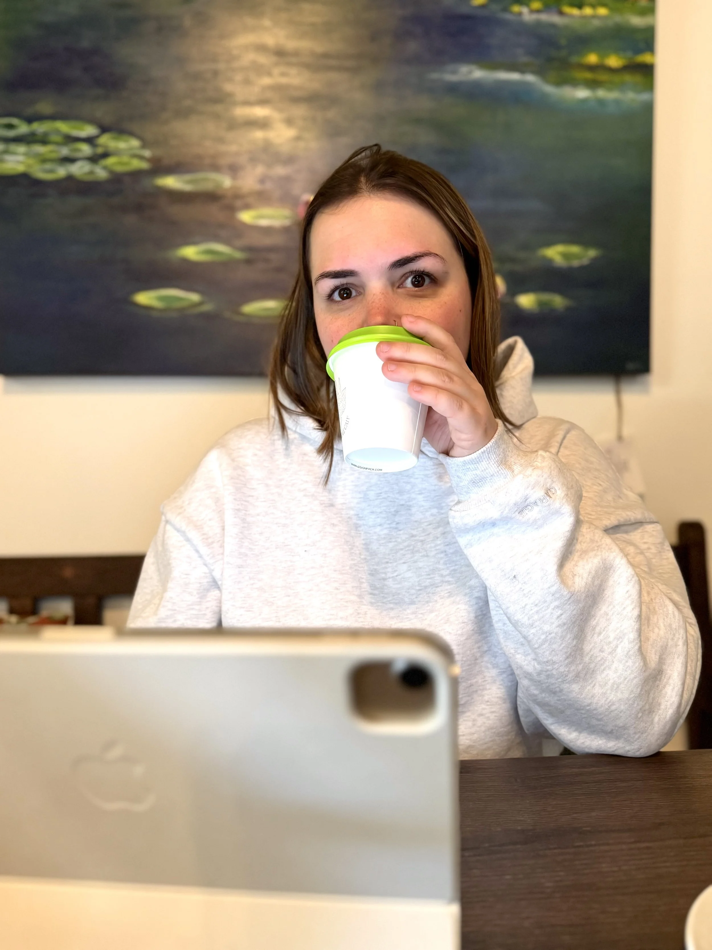 A young woman with brown hair and brown eyes drinking from a white cup with a green lid, sitting at a table in front of a plant-themed painting, with a blurred phone in the foreground.