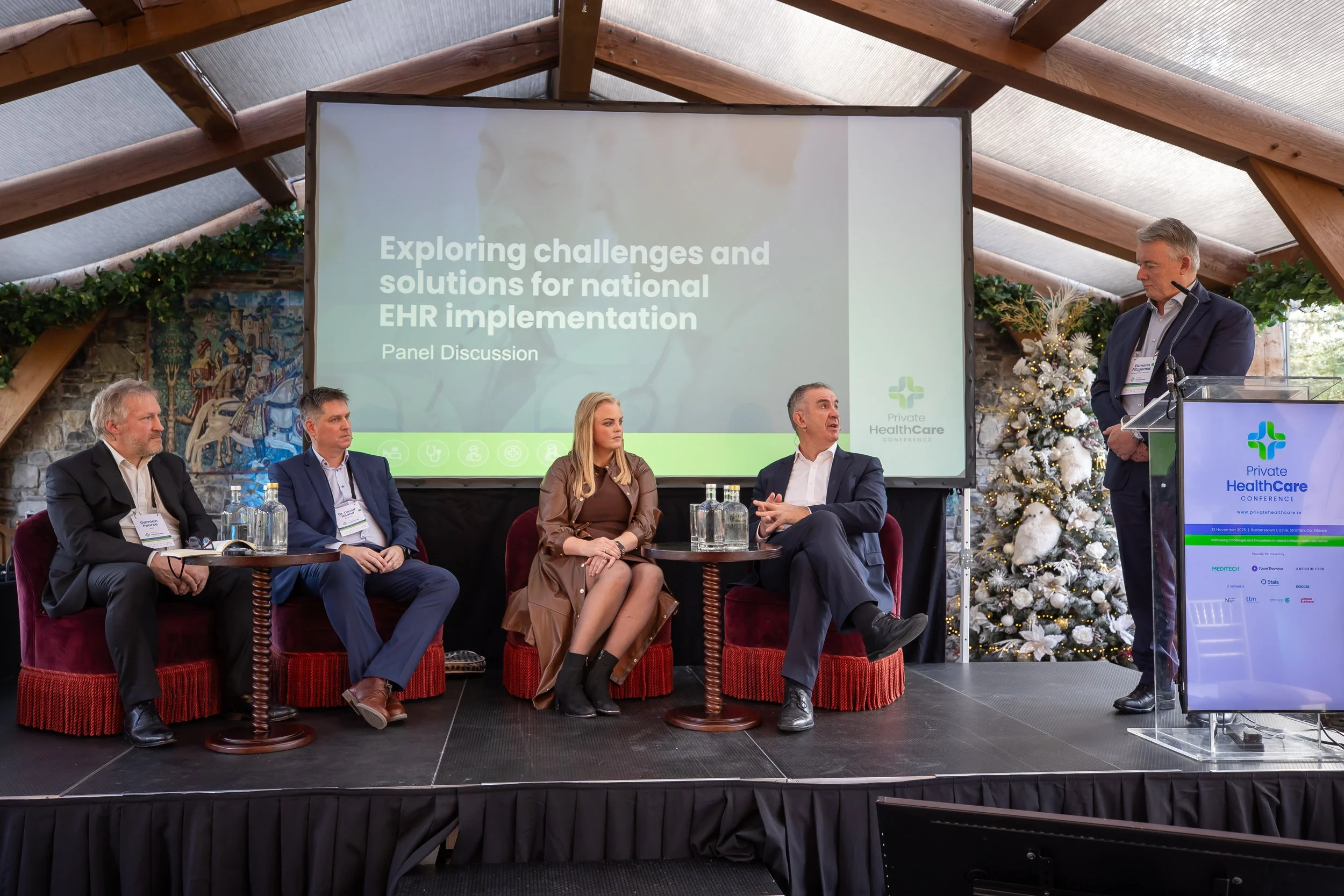 Panel discussion at the Private Healthcare Conference with five professionals seated on stage and a man standing at a podium, in a venue decorated with a Christmas tree and holiday decorations.