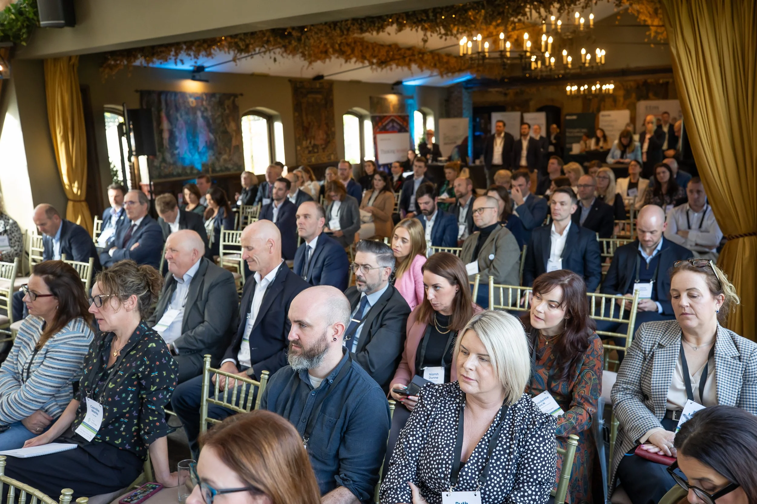 A large group of people sitting in rows at a conference or seminar in a decorated room with chandeliers and paintings, some using tablets or phones.