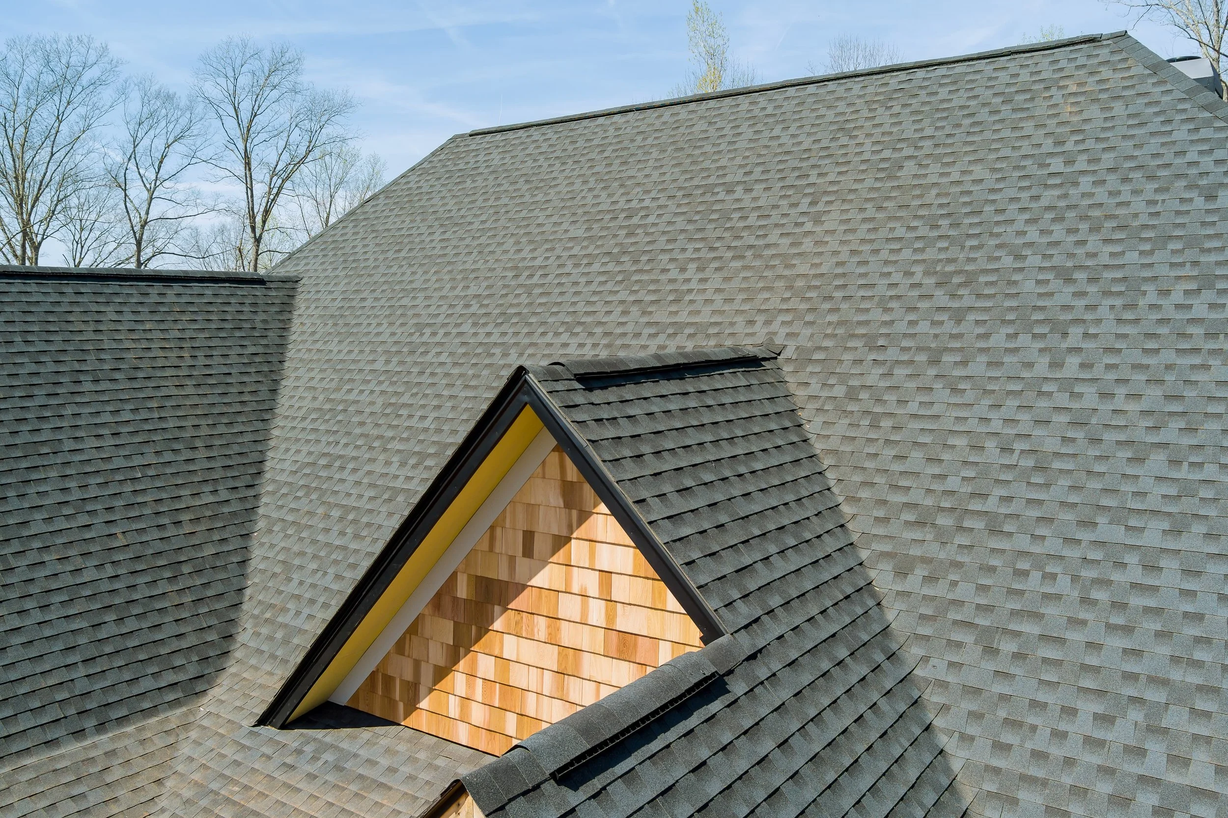 Close-up view of residential rooftops with asphalt shingles and a wooden front porch in the foreground, under a partly cloudy sky with leafless trees in the background.