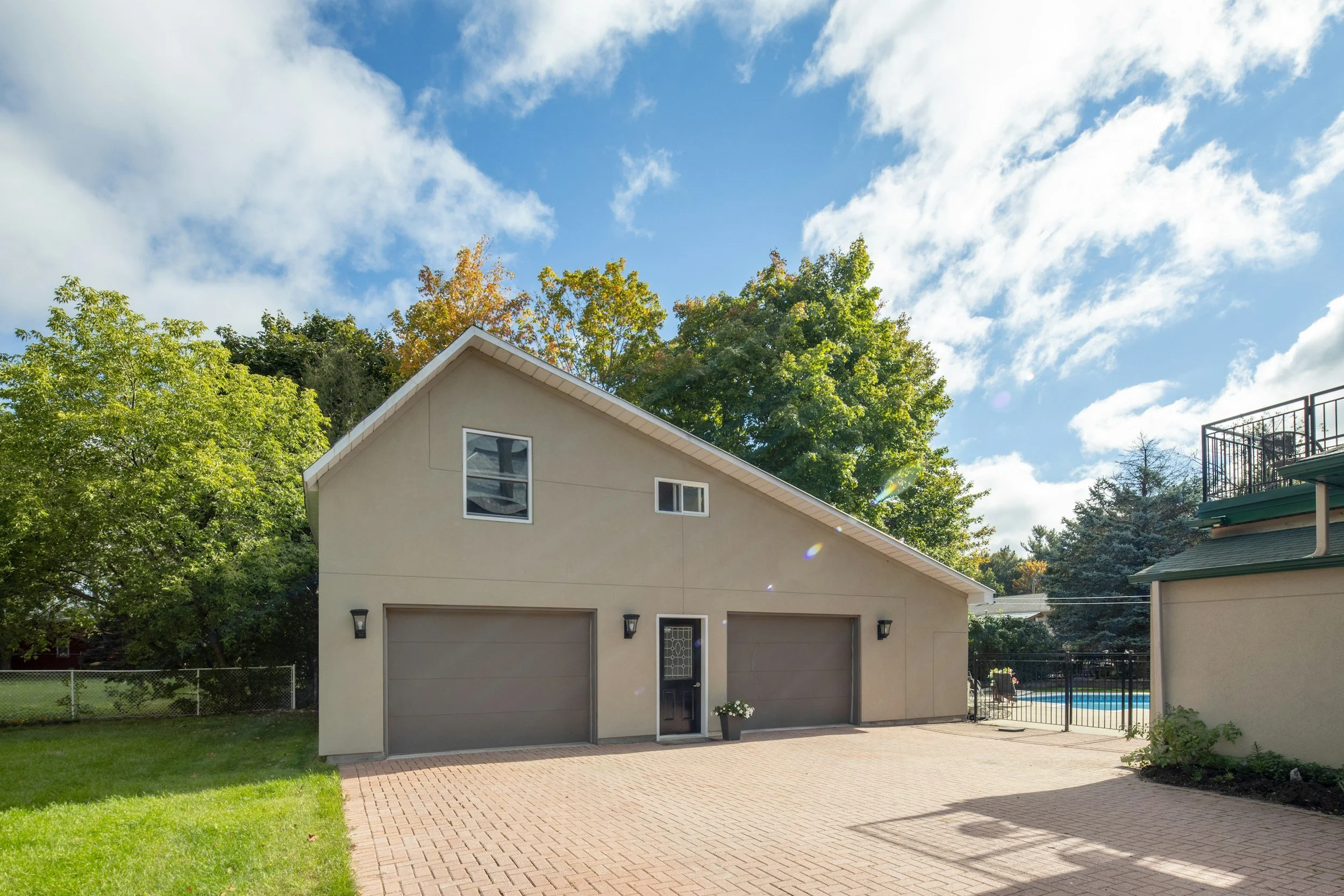 Photo of a beige house with a garage door, side door, and small windows, surrounded by a lawn and tall trees with green and autumn-colored leaves; clear blue sky with some white clouds.