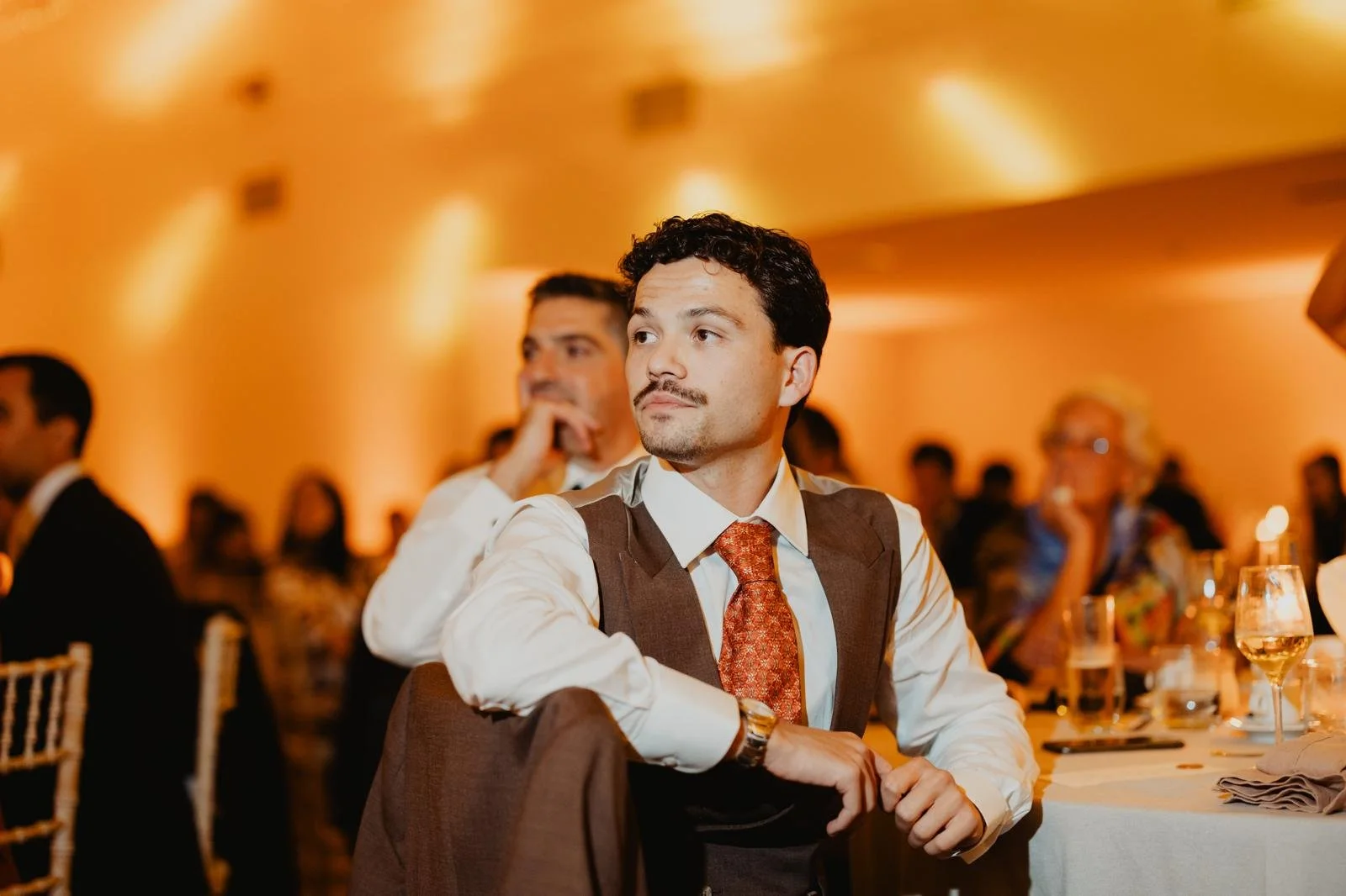 Man sitting at a formal event, dressed in a white shirt, brown vest, and red tie, looking to the side with a serious expression. Several people are in the background, some with glasses on, in a warmly lit room with orange lighting and table settings.
