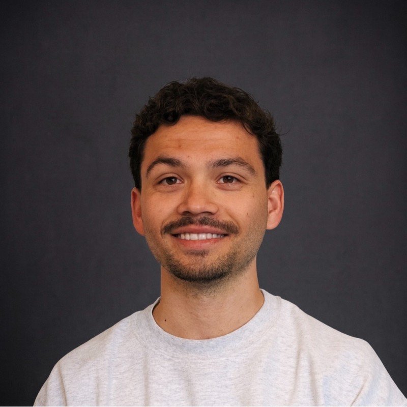 A smiling man with dark curly hair and facial hair, wearing a light gray t-shirt, in front of a dark background.