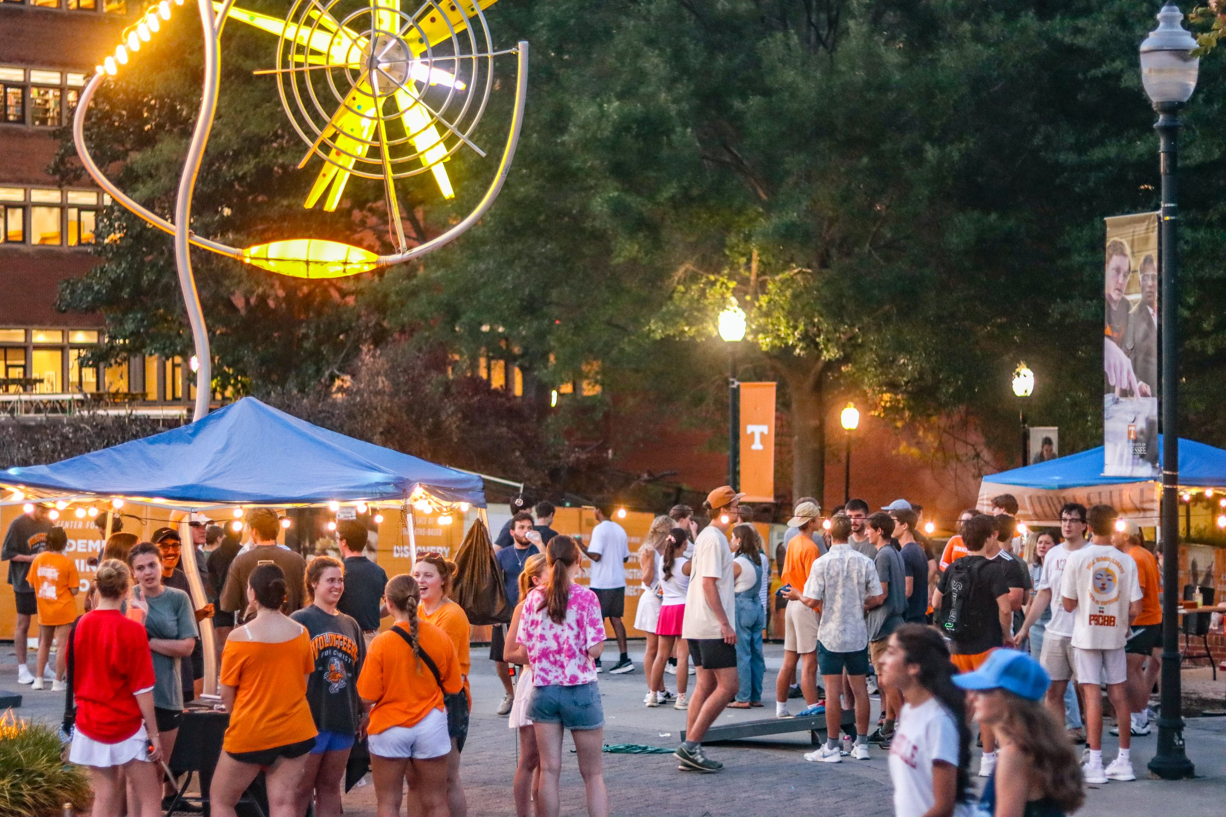 A crowded outdoor event in the evening with people mingling under blue tents and string lights, and a large yellow and black Ferris wheel illuminated in the background amidst green trees and lampposts.