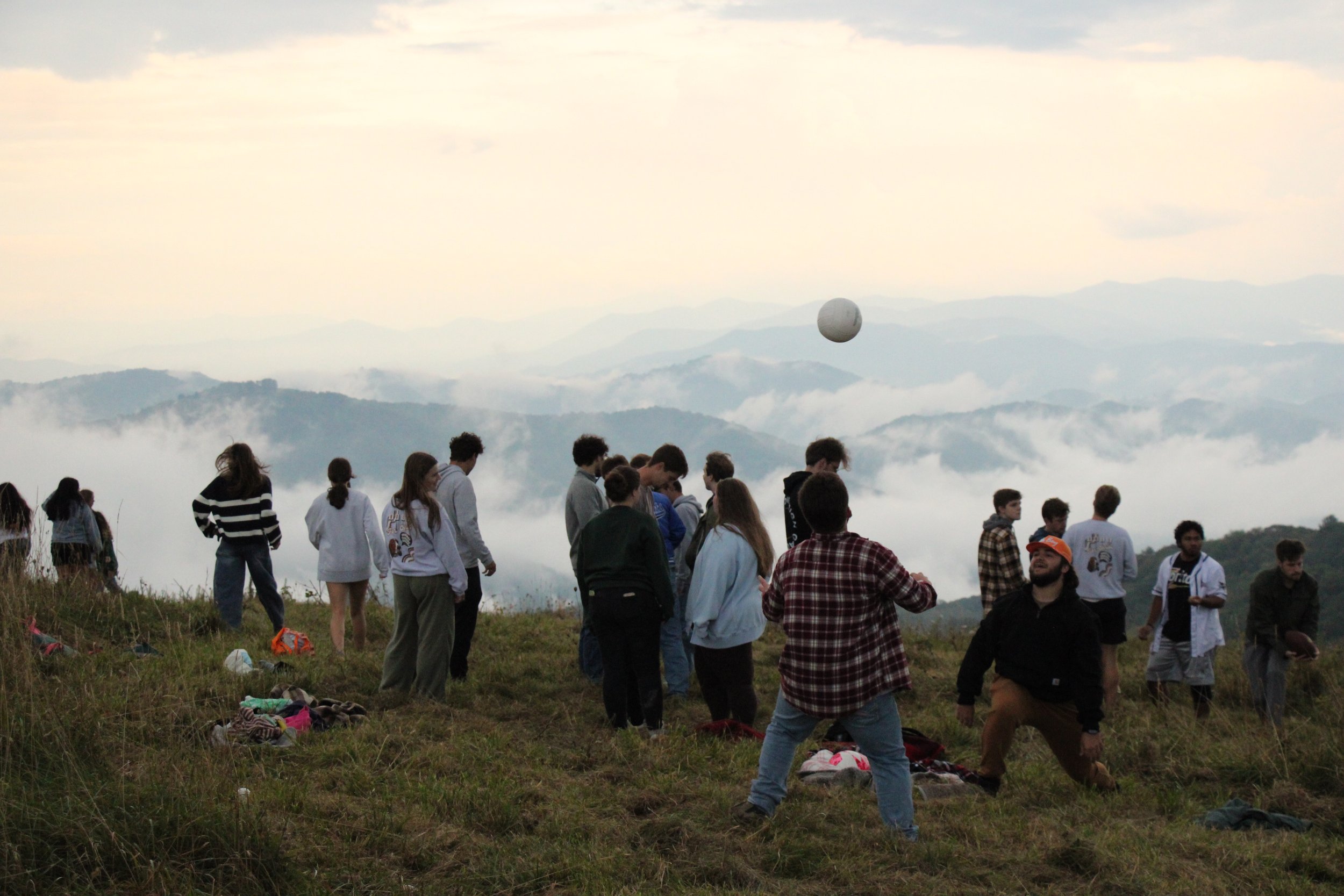 A group of people gathered outdoors on a grassy hillside, watching a large white spherical object float in the sky against a backdrop of mountains and cloudy sky.