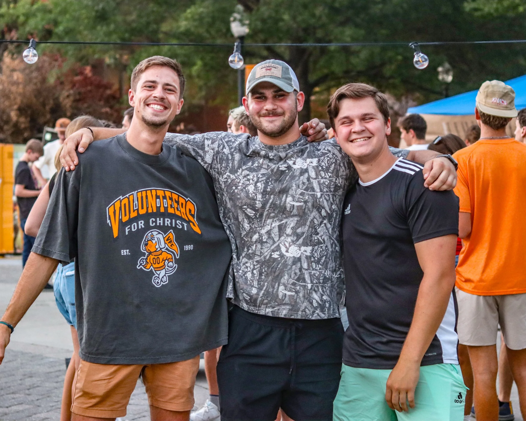 Three smiling young men standing close together outdoors, arms around each other, at an event with other people and string lights in the background.