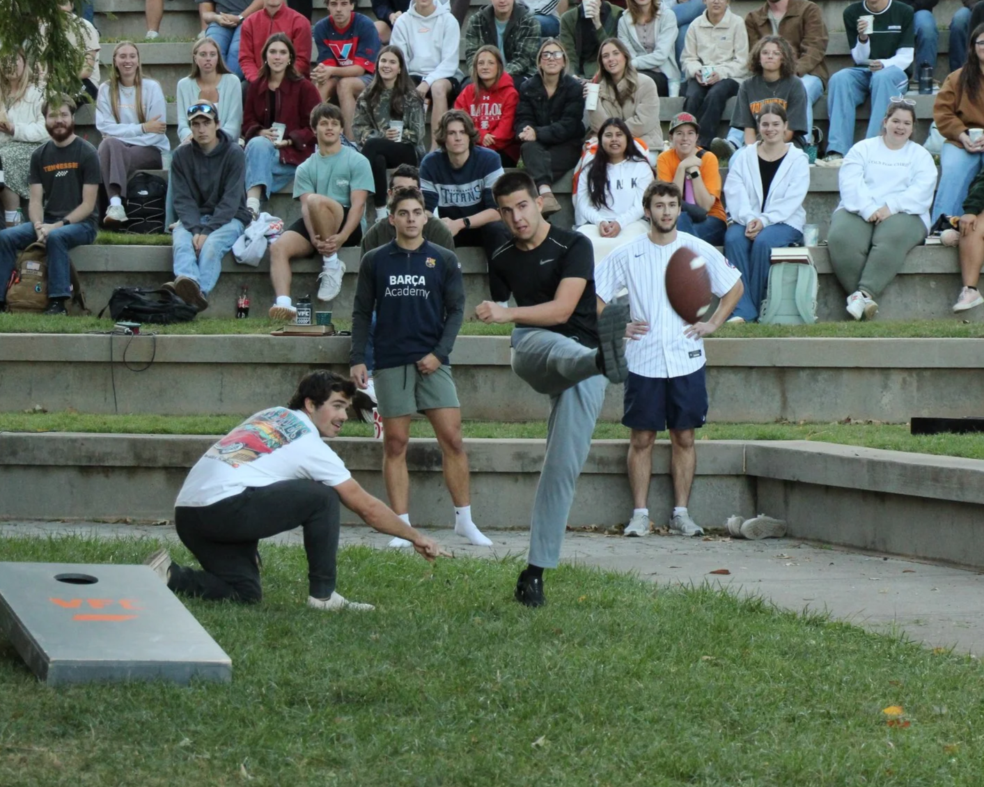 Young men playing a game of cornhole outdoors with a crowd of spectators sitting on tiered concrete steps behind them.
