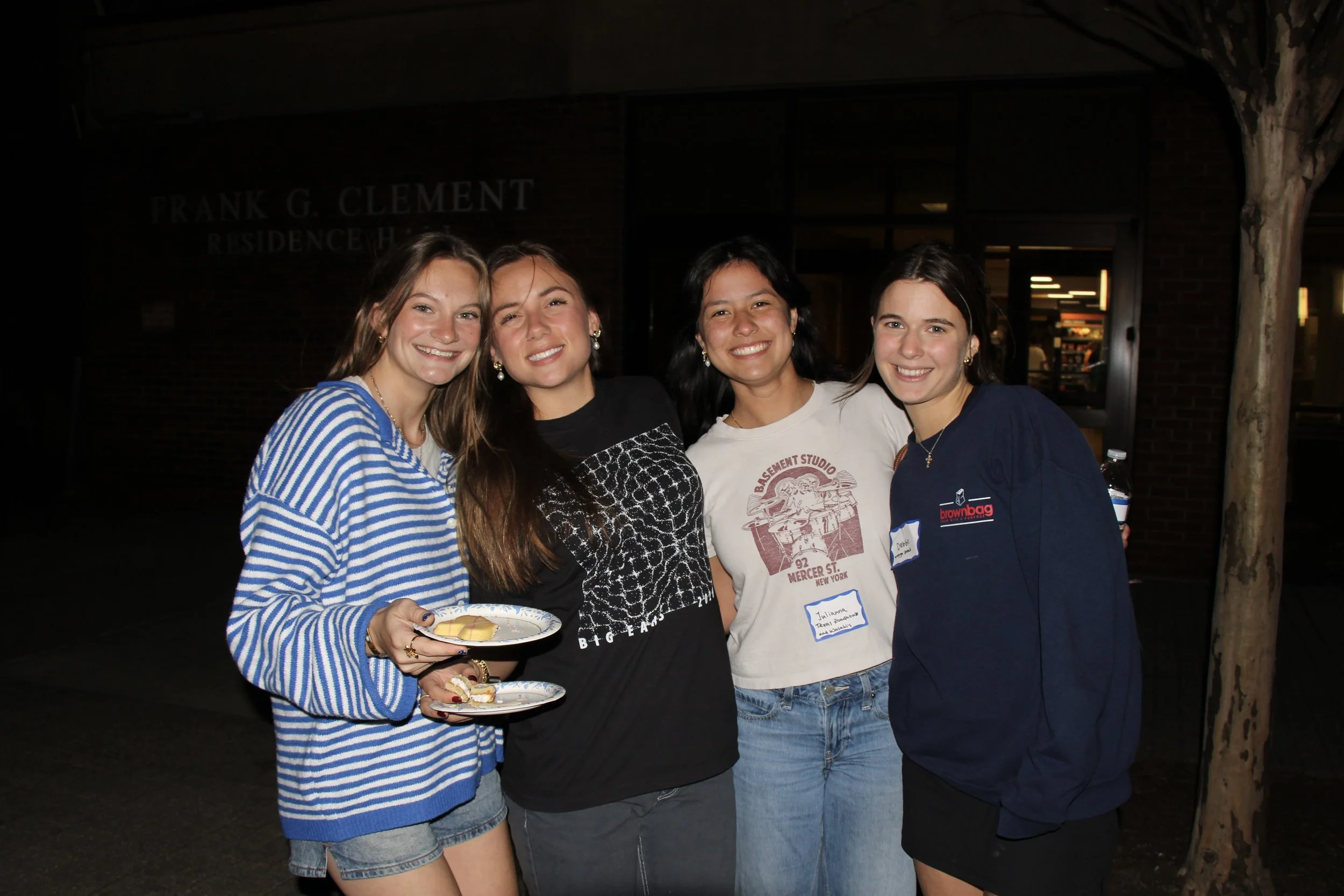 Four women smiling and standing together at night outside a building, with one holding a plate of food.