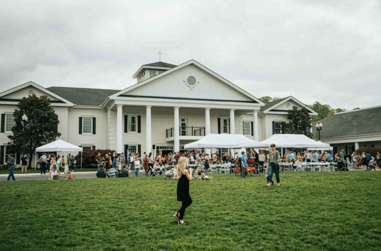 A large crowd gathered outside a white church with columns, under tents, for an event on a cloudy day, with people sitting, walking, and standing on the grass and near the building.
