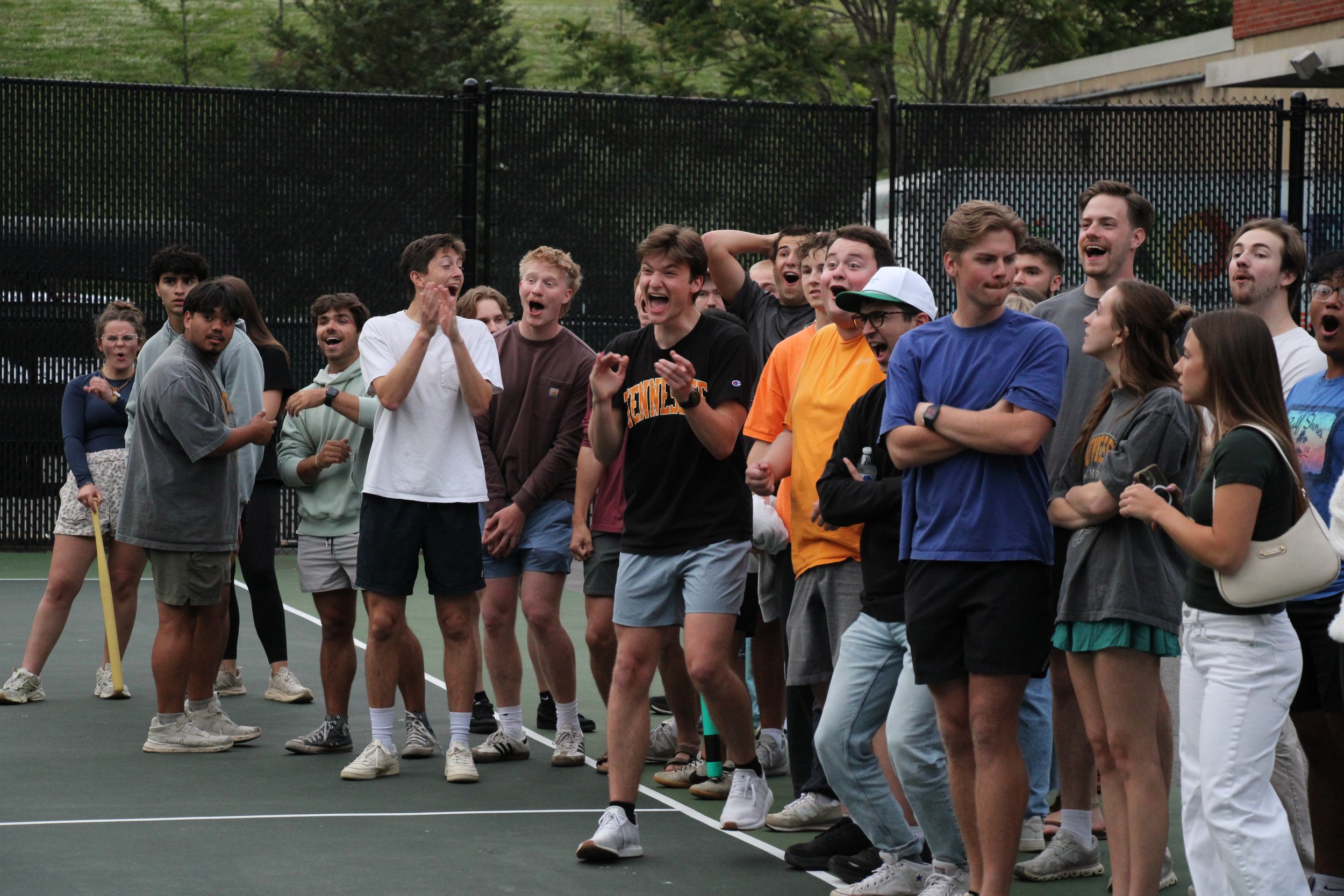 Group of people gathered on an outdoor tennis court, looking excited and happy, with a black fence and trees in the background.