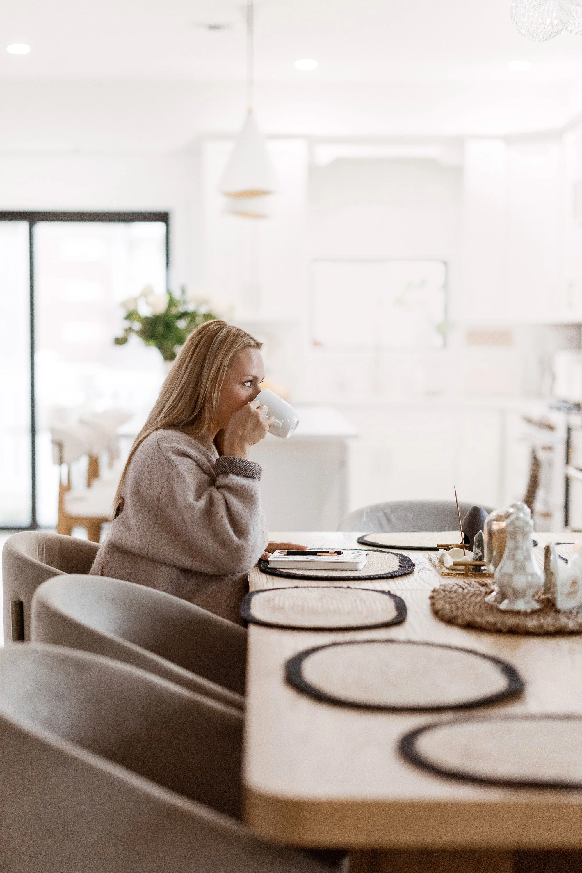 A woman with long blonde hair sitting at a dining table, drinking coffee from a white mug, in a bright, modern kitchen with white walls and natural light.