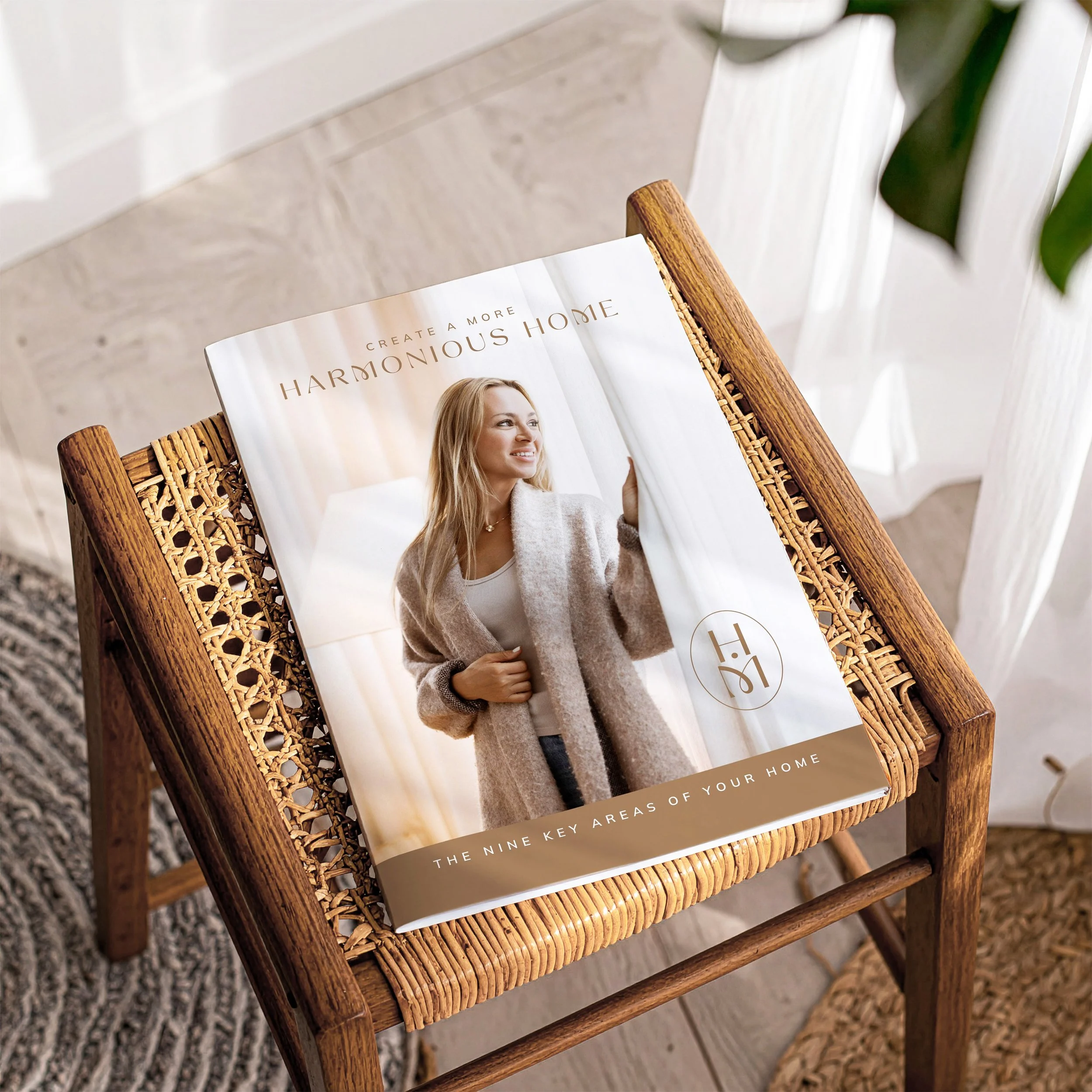 A wicker chair with a magazine titled 'Create a More Harmonious Home' with a woman on the cover, placed on a beige rug near a white wall and green plant leaves.
