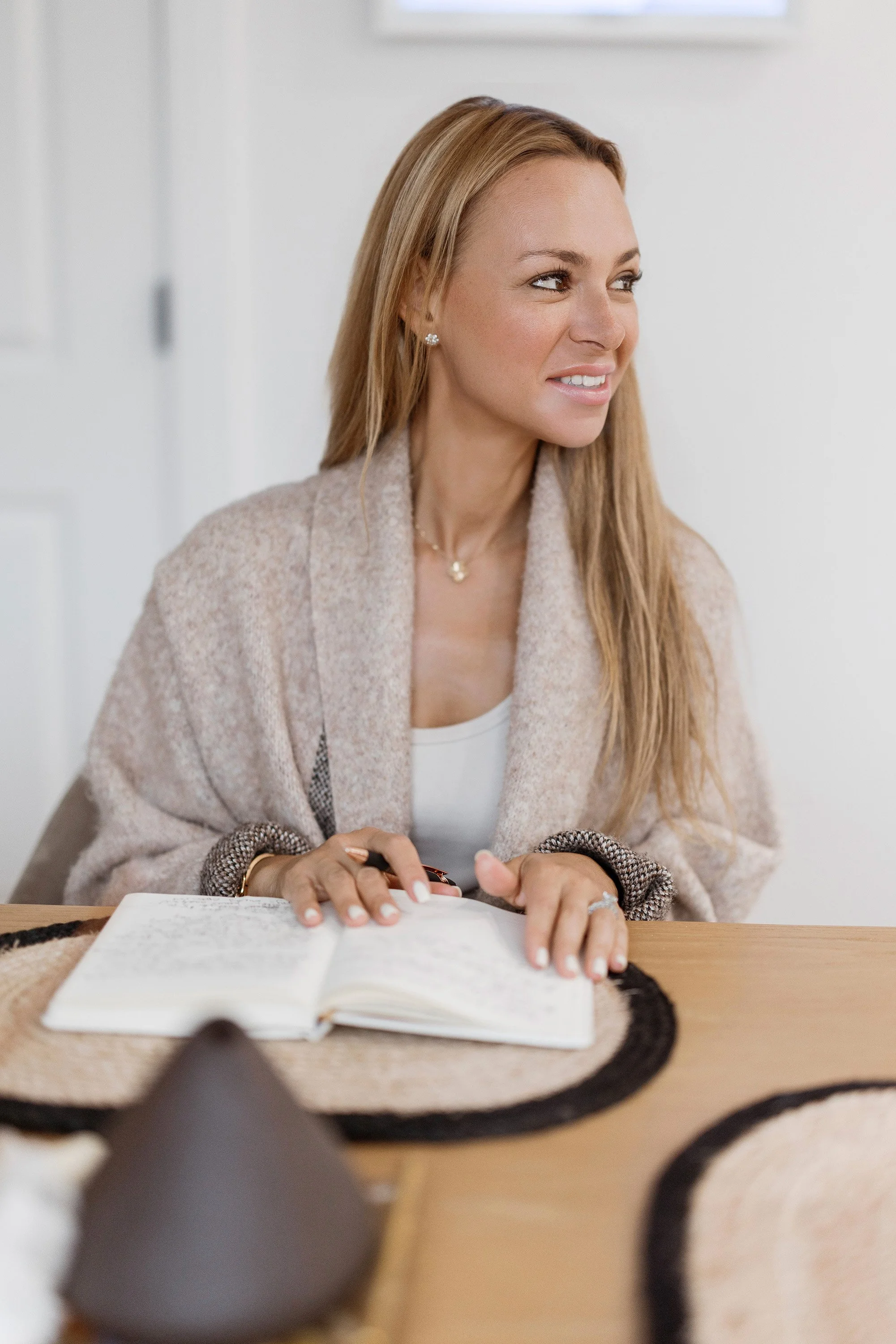 A woman with long blonde hair, light-colored sweater, and jewelry sits at a table with an open notebook, smiling and looking to her left.