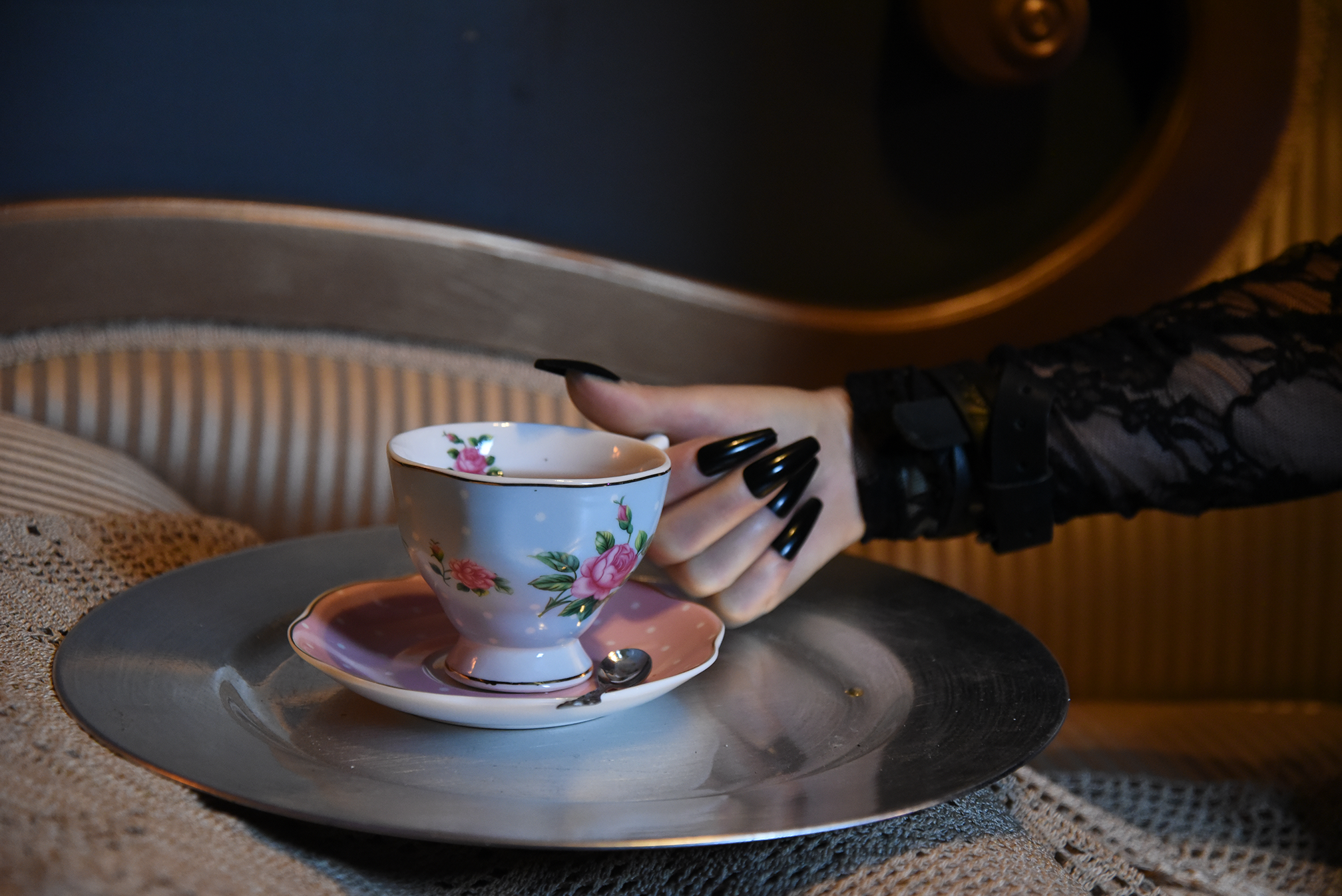 FemDom with black long nails holding a floral teacup and saucer, positioned over a silver serving tray on a textured surface, with lace fabric and chaise lounge.