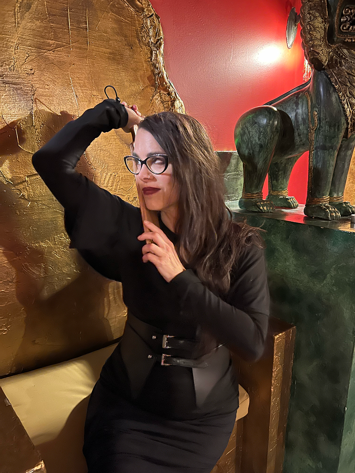 A woman with long dark hair and glasses striking a playful pose with her hand on a paddle, sitting in front of a golden textured wall and an ornate sculpture of an animal with green and black colors.