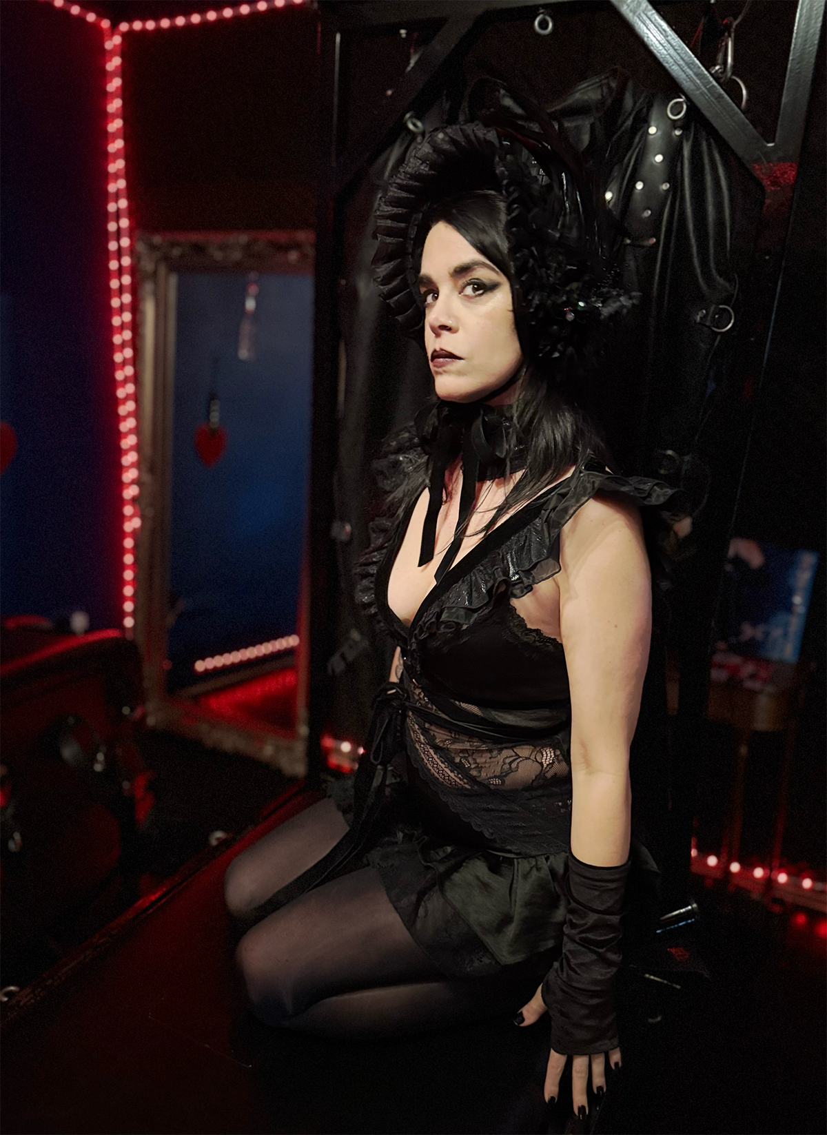 A woman with dark hair dressed in black lingerie, stockings, and gloves, sitting in a dark, gothic setting with red lighting and heart-shaped decorations in the background.