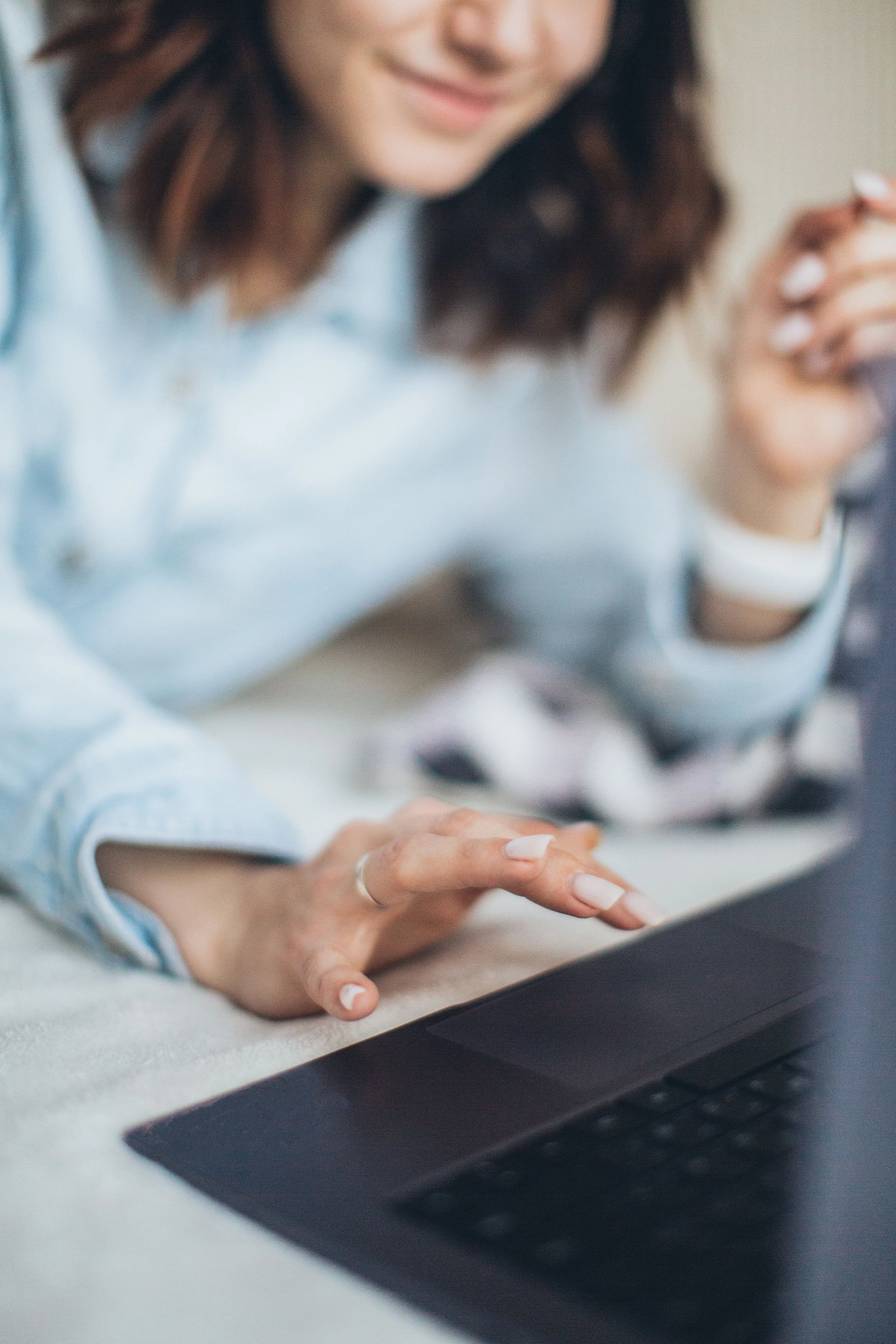 Close-up of a woman's hand using a laptop keyboard, wearing a ring, with a blurred woman in the background smiling, sitting at a desk.