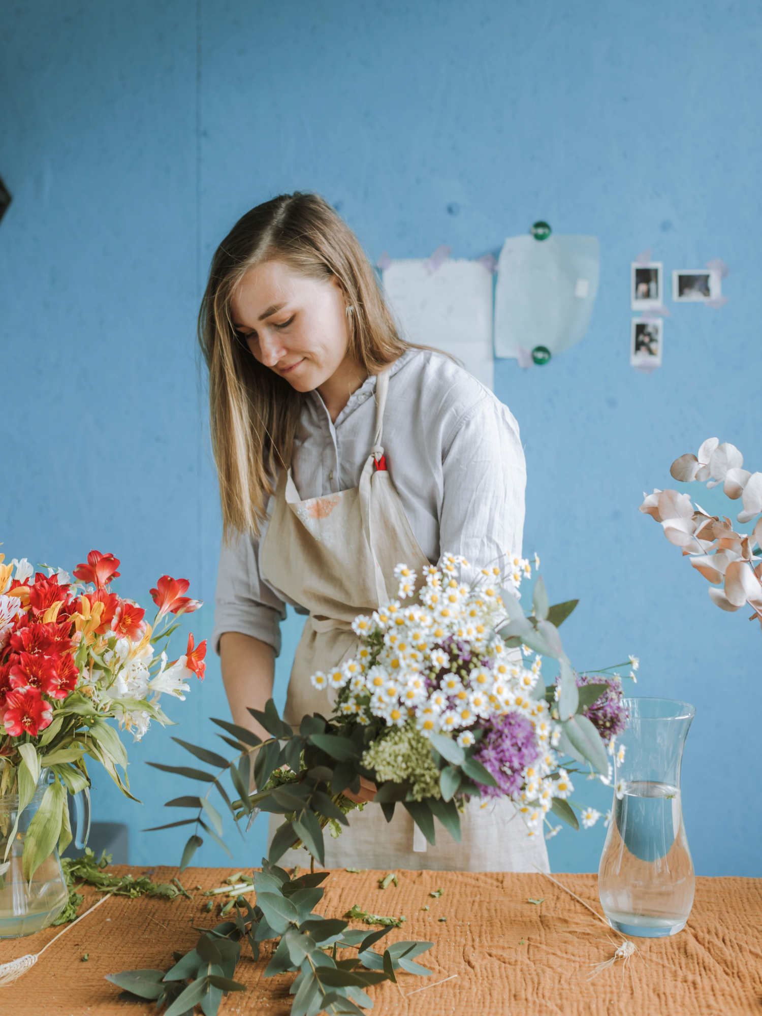 A florist arranging a flower bouquet at a table with other floral arrangements, against a blue wall with pinned paper and photos.