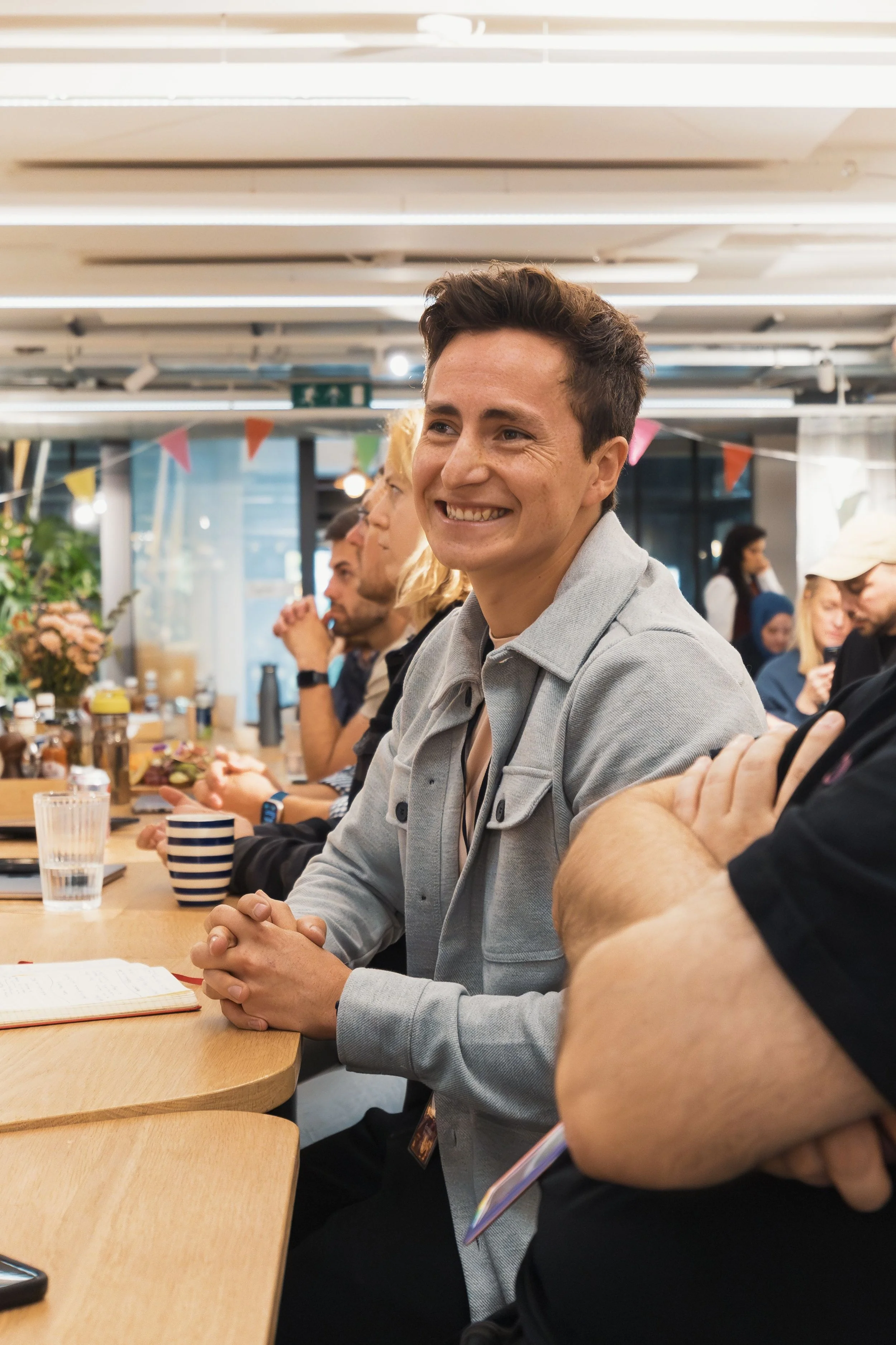 A group of people sitting at a wooden table in a bright, modern indoor space, with a man smiling and clasping his hands together in the foreground. There are drinks, notebooks, and snacks on the table, and colorful bunting hanging from the ceiling in the background.