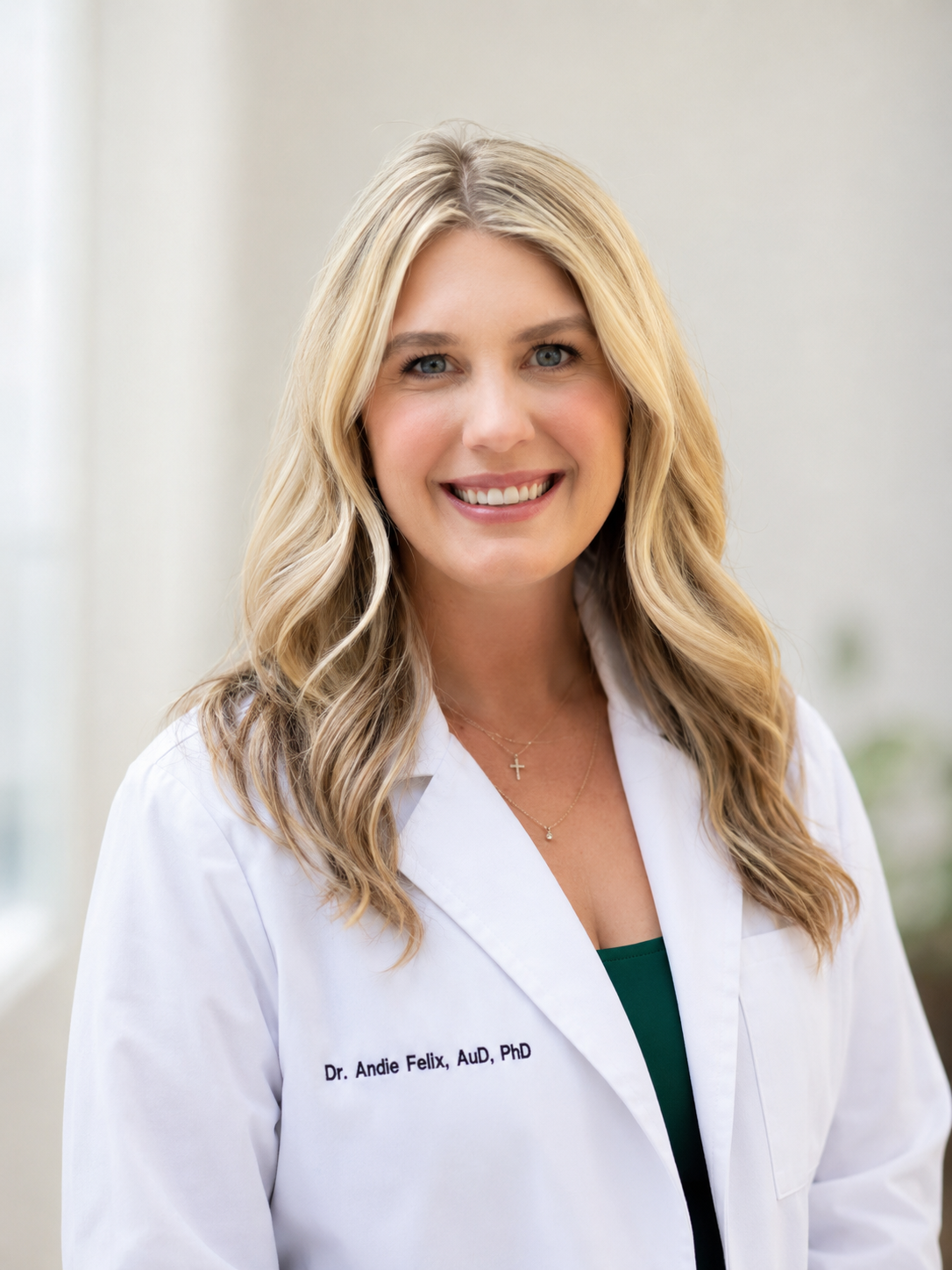 A smiling woman with blonde hair wearing a white lab coat with the name "Dr. Andie Felix, AuD, PhD" embroidered on it, standing indoors.