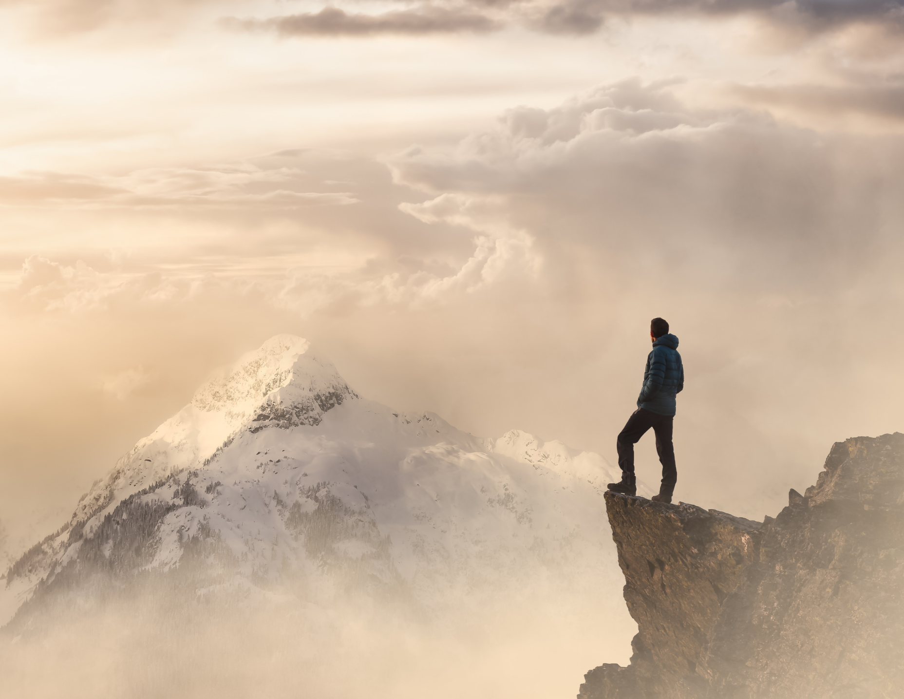A person standing on a rocky ledge with snow-capped mountains and a cloudy sky in the background.
