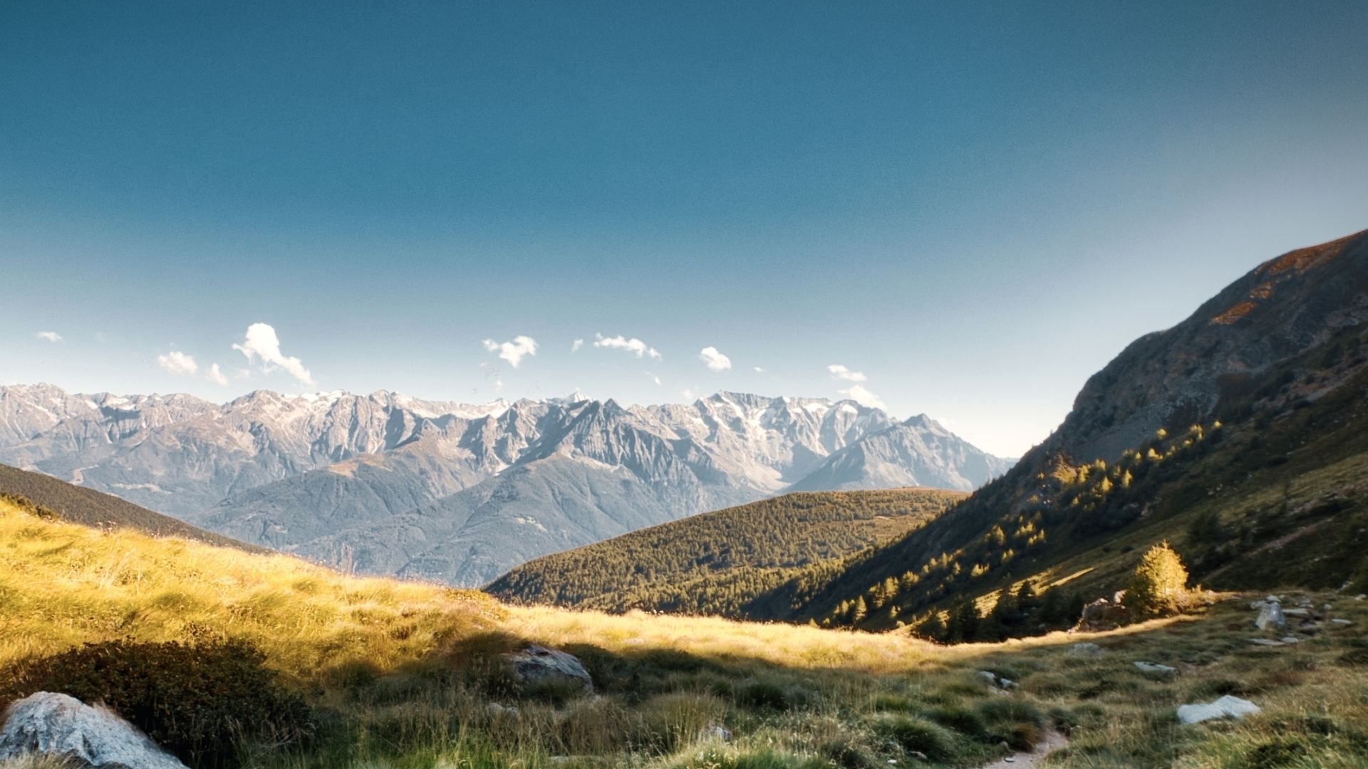 A scenic landscape of a mountain range with snow-capped peaks, green hills, and patches of trees under a clear blue sky with small clouds.