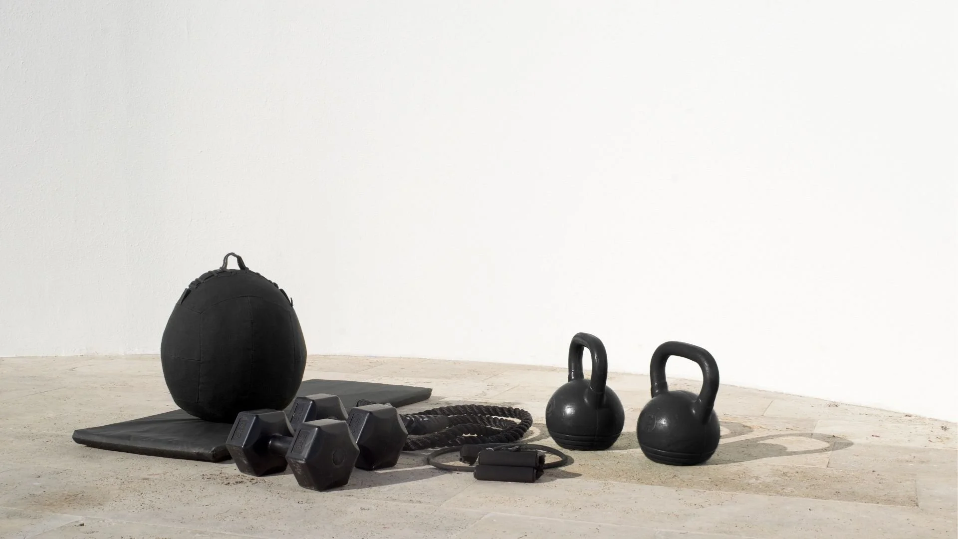 Assorted gym equipment including a medicine ball, kettlebells, dumbbells, a foam roller, a jump rope, and a yoga mat arranged on a concrete floor against a plain white wall