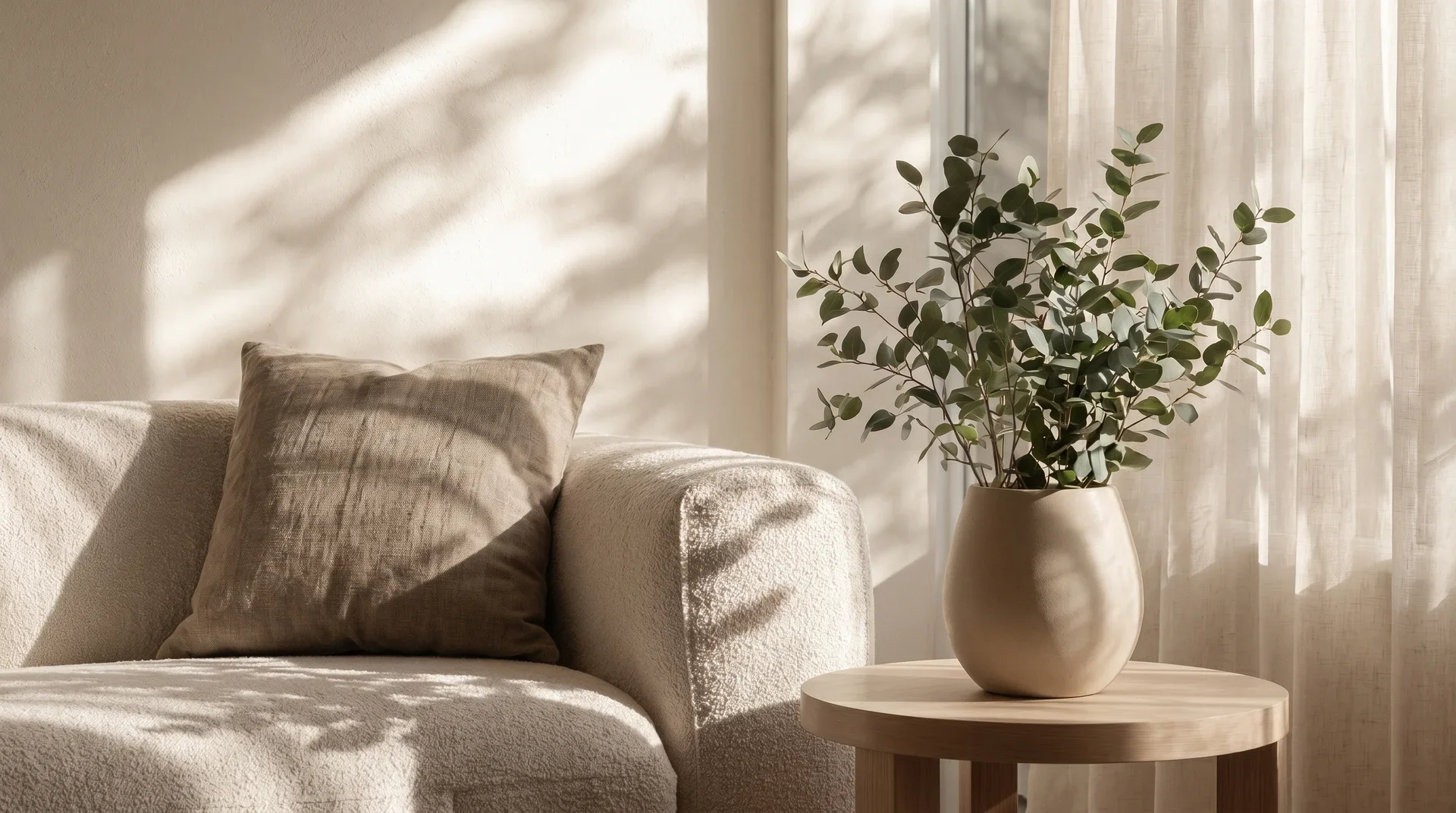 A beige sofa with a thrown pillow, a small wooden round side table with a clay vase, and a houseplant near a window with sheer curtains, illuminated by natural sunlight.