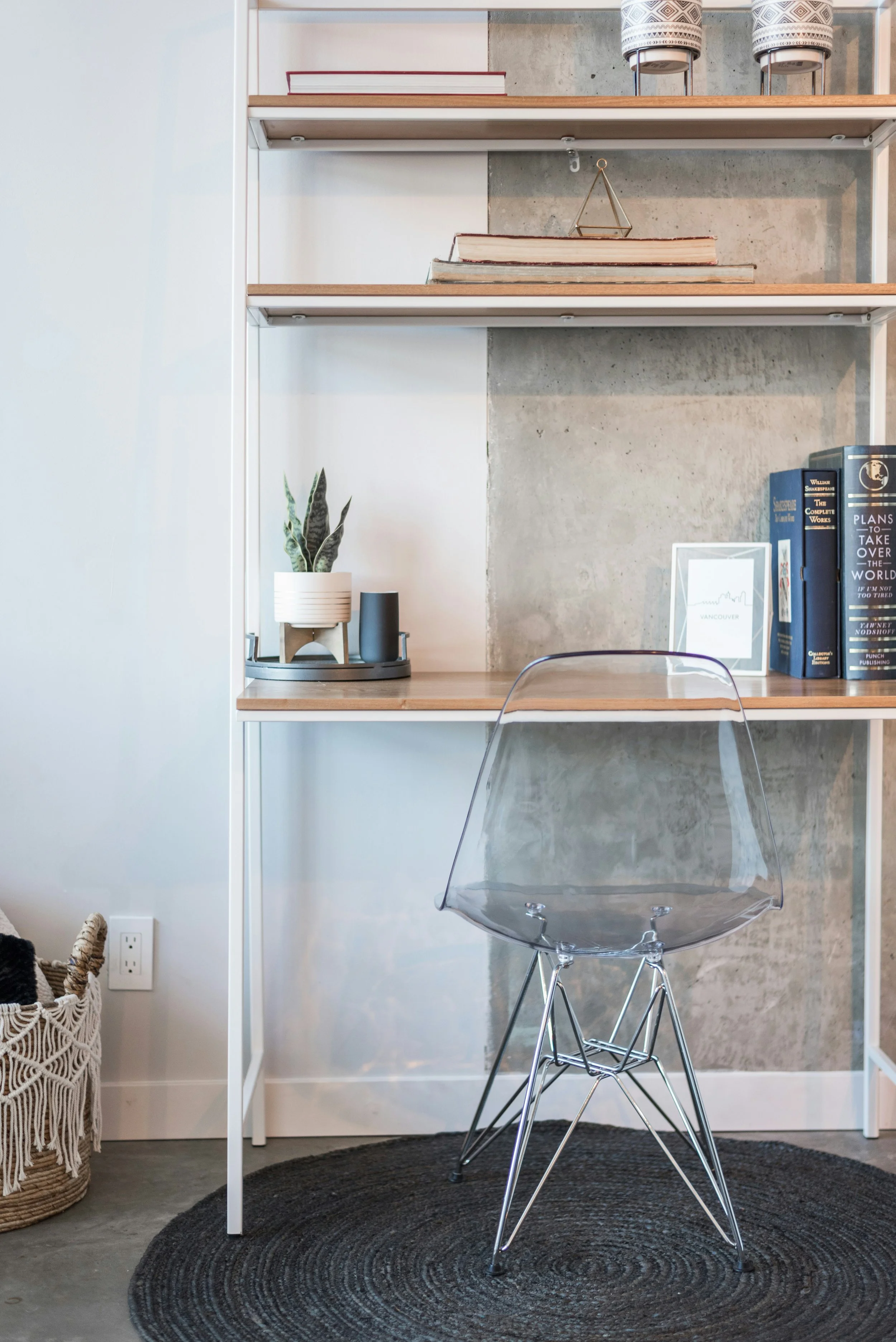 A modern workspace with a clear acrylic chair centered on a round, black braided rug. Behind the chair is a built-in desk with a white metal frame and a light wood top. On the desk, there is a potted snake plant with pointed green leaves, a small dark gray vase, and a white tray. Above the desk, there are three shelves with books, decorative items, and a small framed art print that says 'Vancouver'. The background wall features a concrete and white panel design, with a power outlet near the basket on the floor to the left.