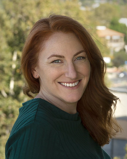 A woman with red hair and a big smile standing outdoors, with trees and buildings in the background.