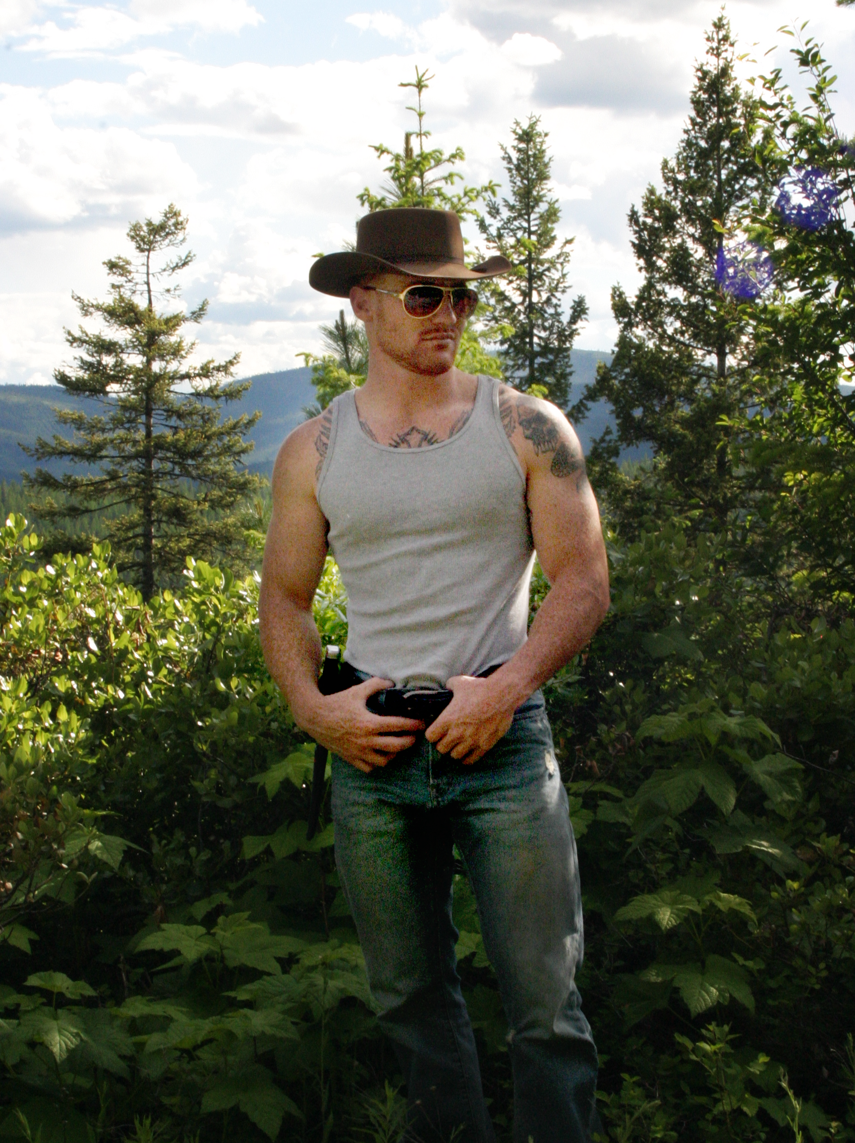 A man wearing a brown cowboy hat, sunglasses, a gray tank top, and jeans standing outdoors among green plants and trees with a mountainous landscape and partly cloudy sky in the background.