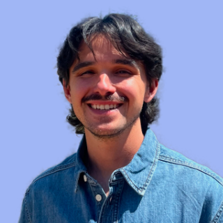 A smiling young man with dark hair and a beard, wearing a denim shirt against a blue sky background.