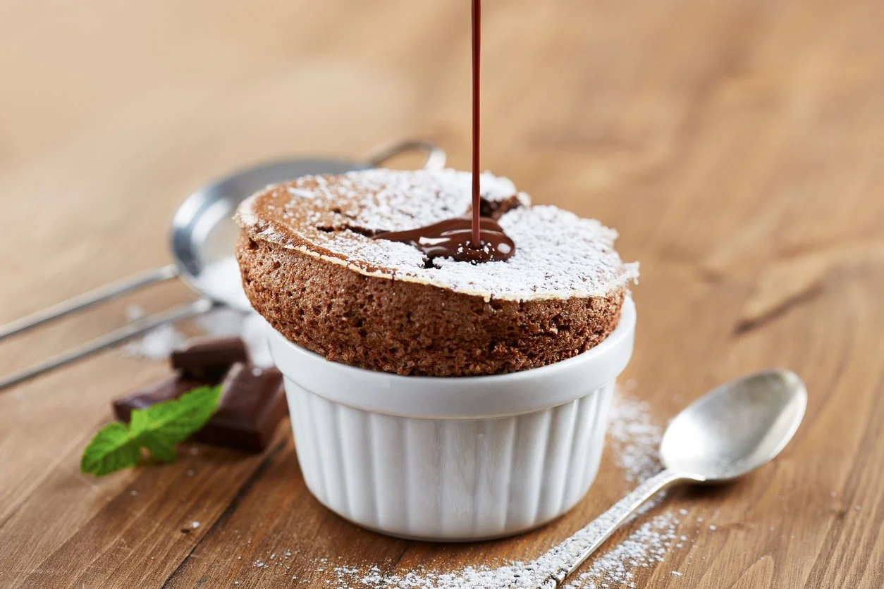 Chocolate lava cake with powdered sugar being topped with chocolate syrup in a white ramekin on a wooden surface, with mint leaves and chocolate pieces nearby.