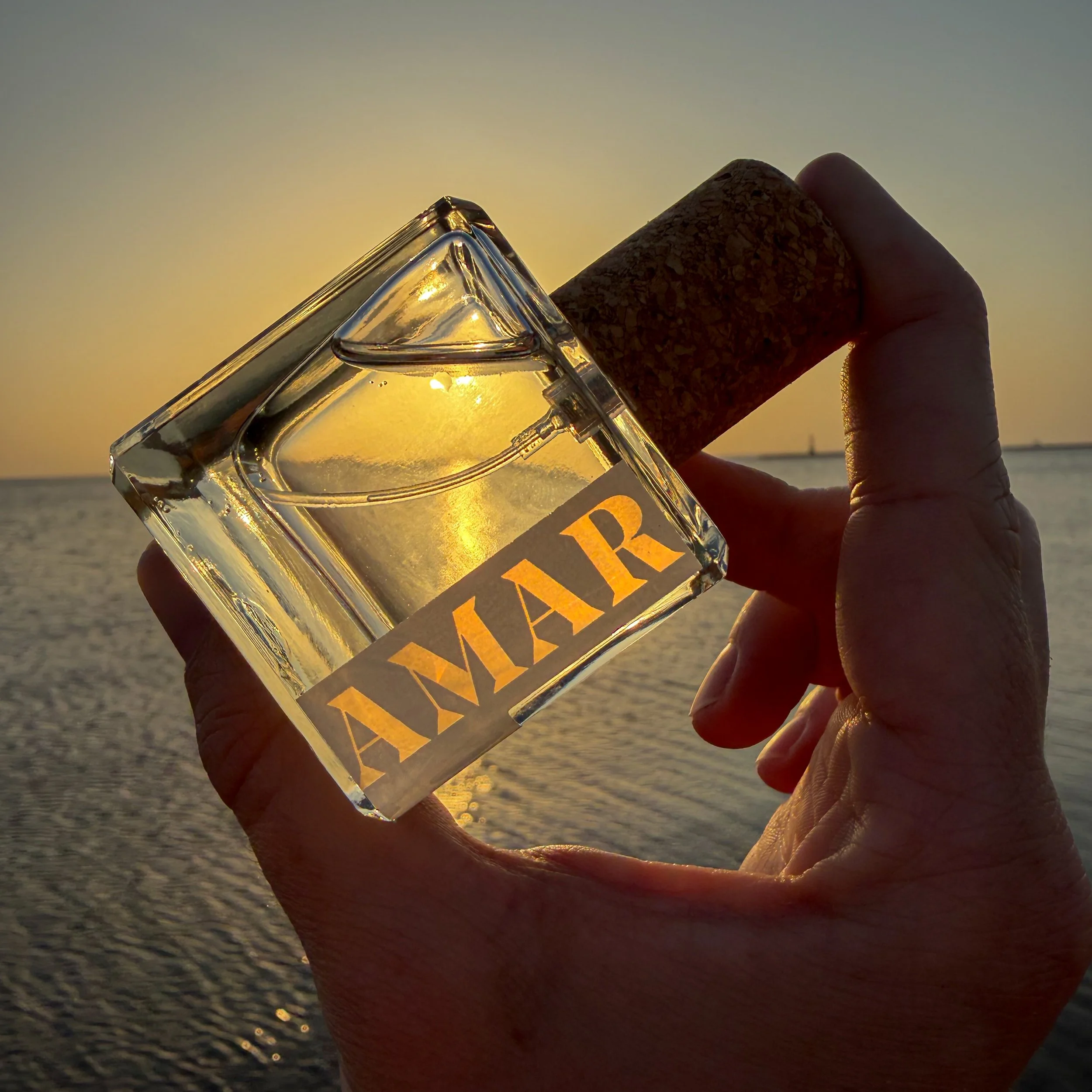 Close-up of a hand holding a glass perfume bottle and a cork stopper against a sunset background by the water.