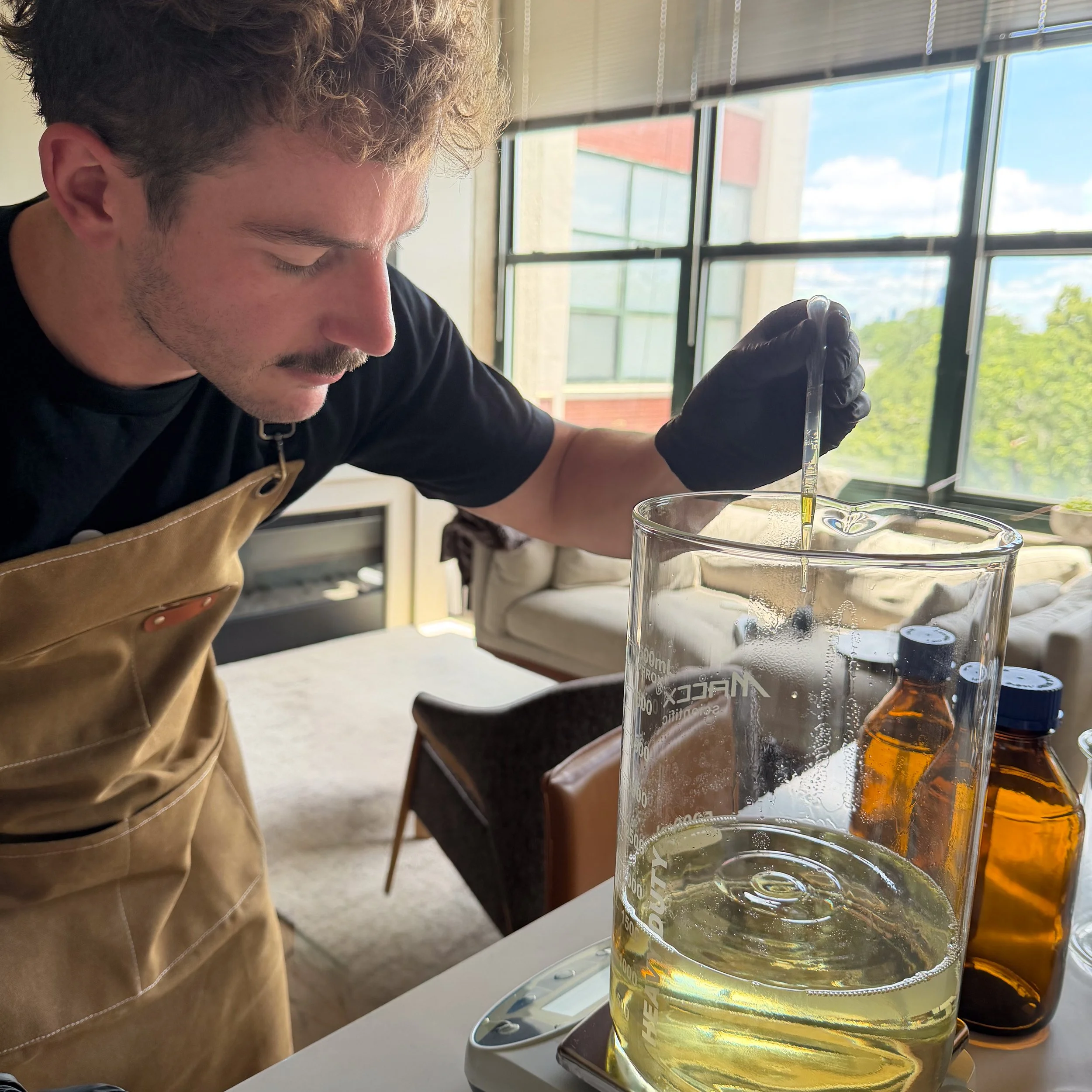 A person with curly hair, wearing a black shirt, gloves, and an apron, is conducting a science experiment with liquids in a lab or classroom with large windows showing a bright day outside.