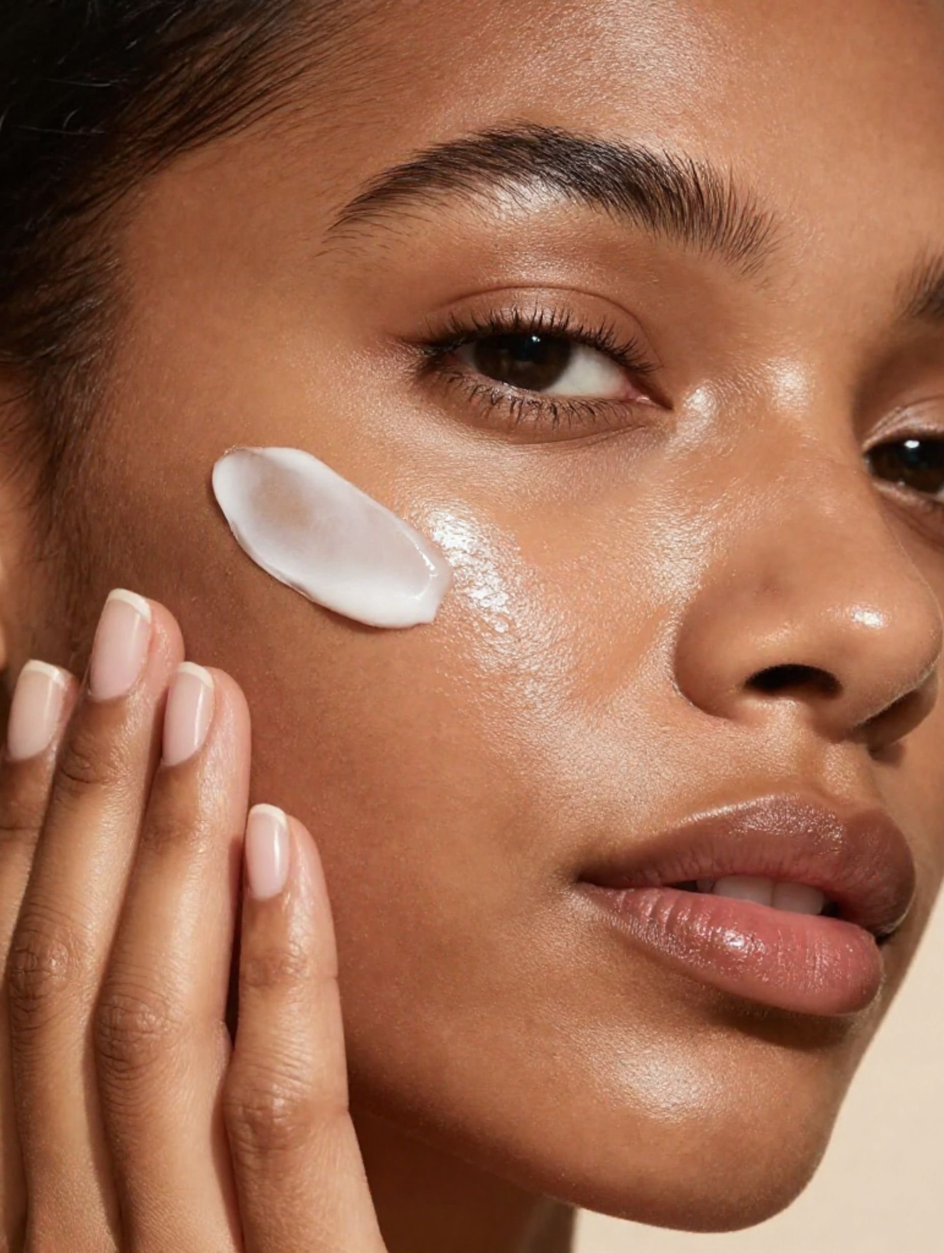A close-up of a woman's face with fair skin applying a white skincare cream to her cheek with her fingers, focusing on her eye and cheek area.