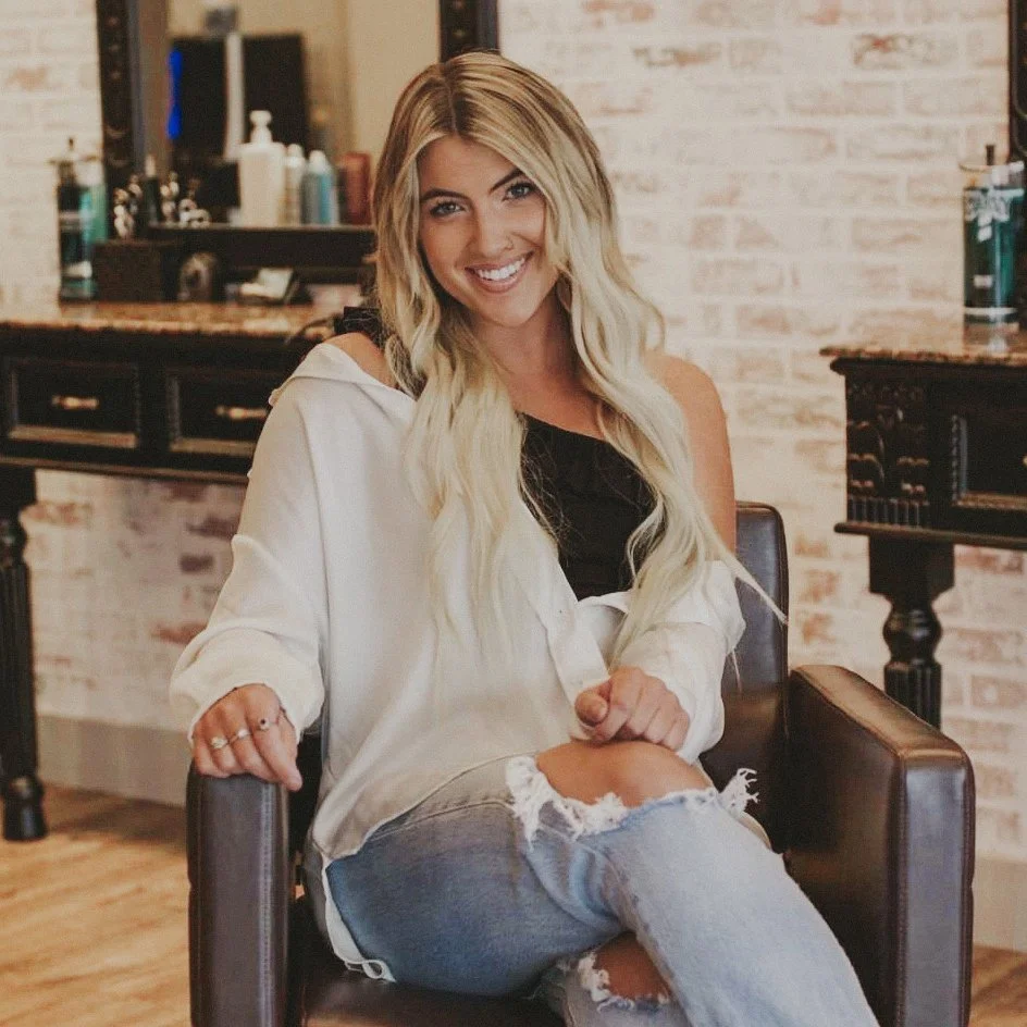 A young woman with long blonde hair, smiling, sitting in a hair salon chair, wearing a white shirt and ripped jeans.