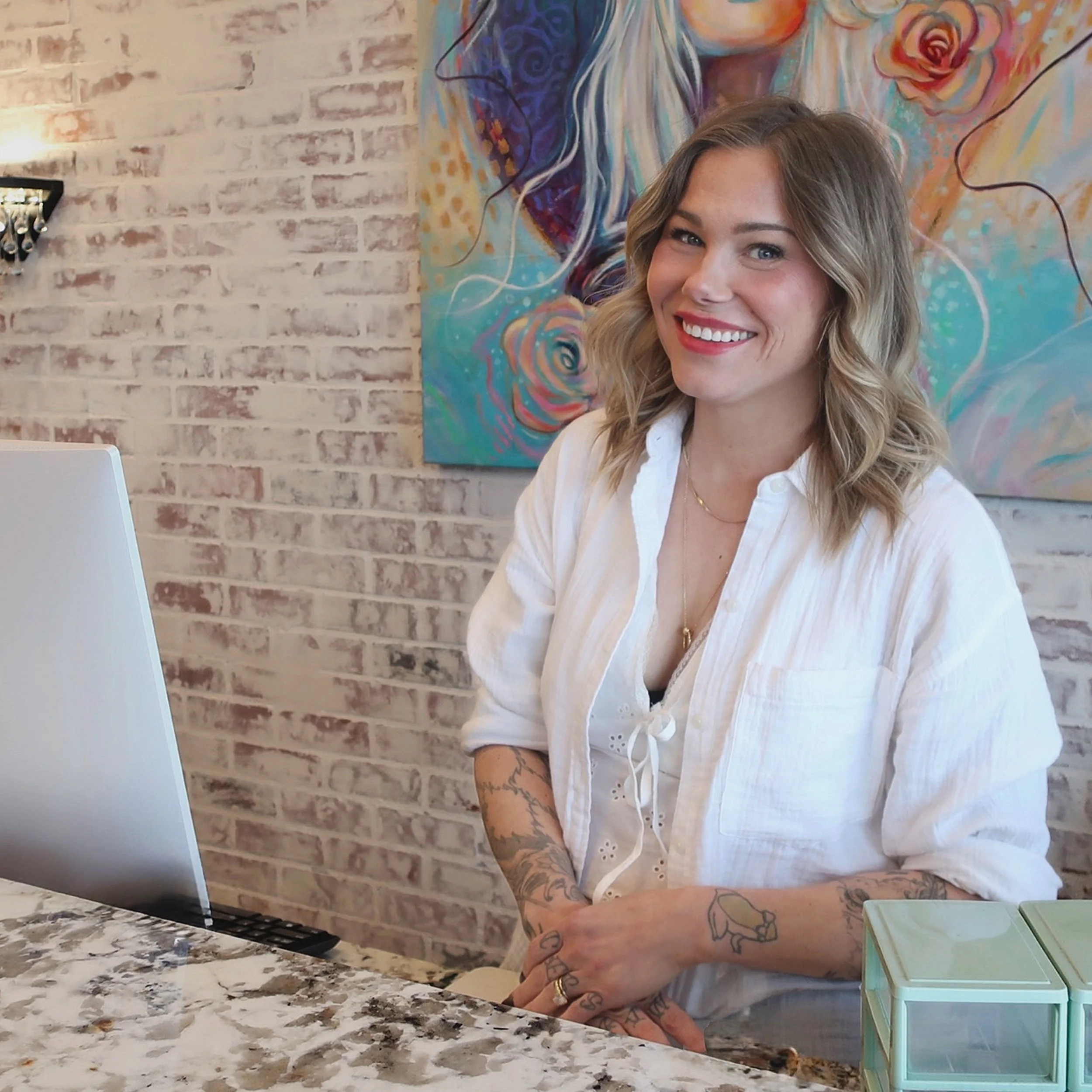 A woman with wavy blonde hair and tattoos on her arms is smiling while sitting at a marble countertop in a cafe. Behind her, a painted wall features a colorful abstract portrait of a woman with roses in her hair. The cafe has a white brick wall and a black wall sconce with candles.