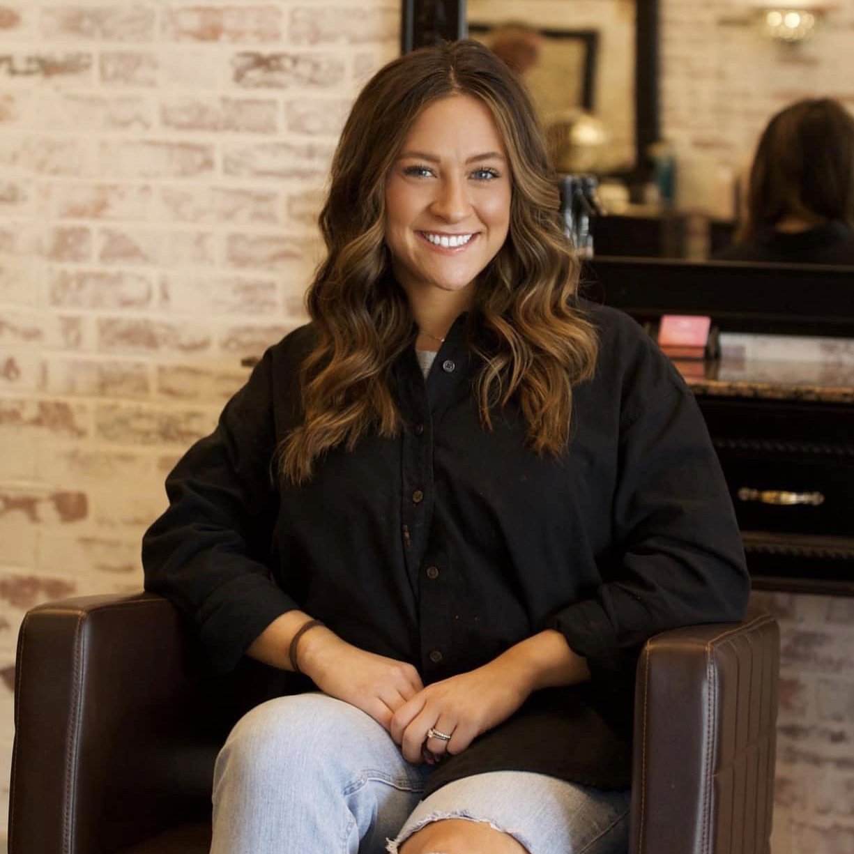 A woman with long, wavy brown hair, smiling, sitting in a brown leather chair in a cozy, brick-walled room, wearing a black button-up shirt and light jeans.