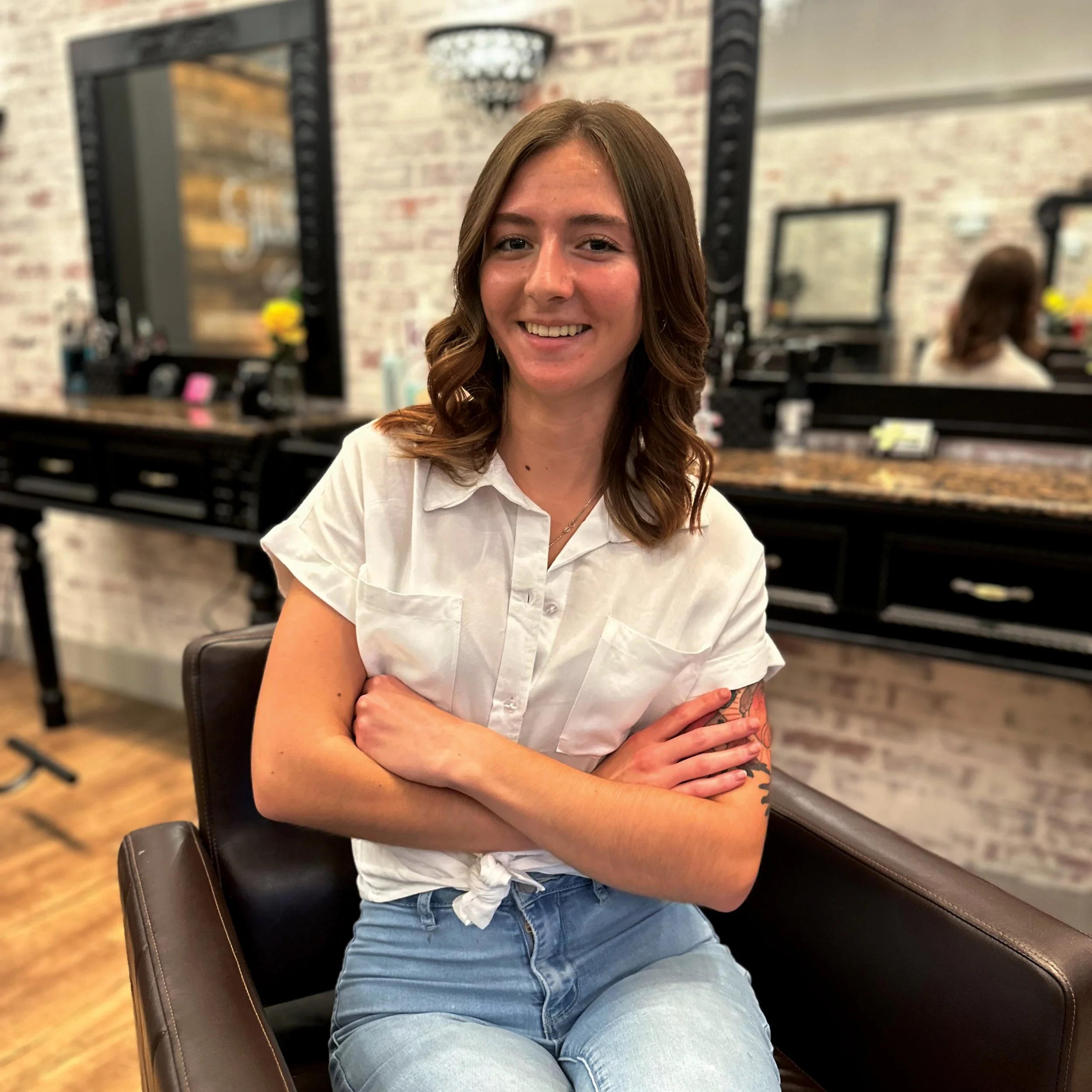 A smiling woman with brown hair and a white shirt sitting in a salon or barbershop with mirrors, hair products, and a brick wall in the background.