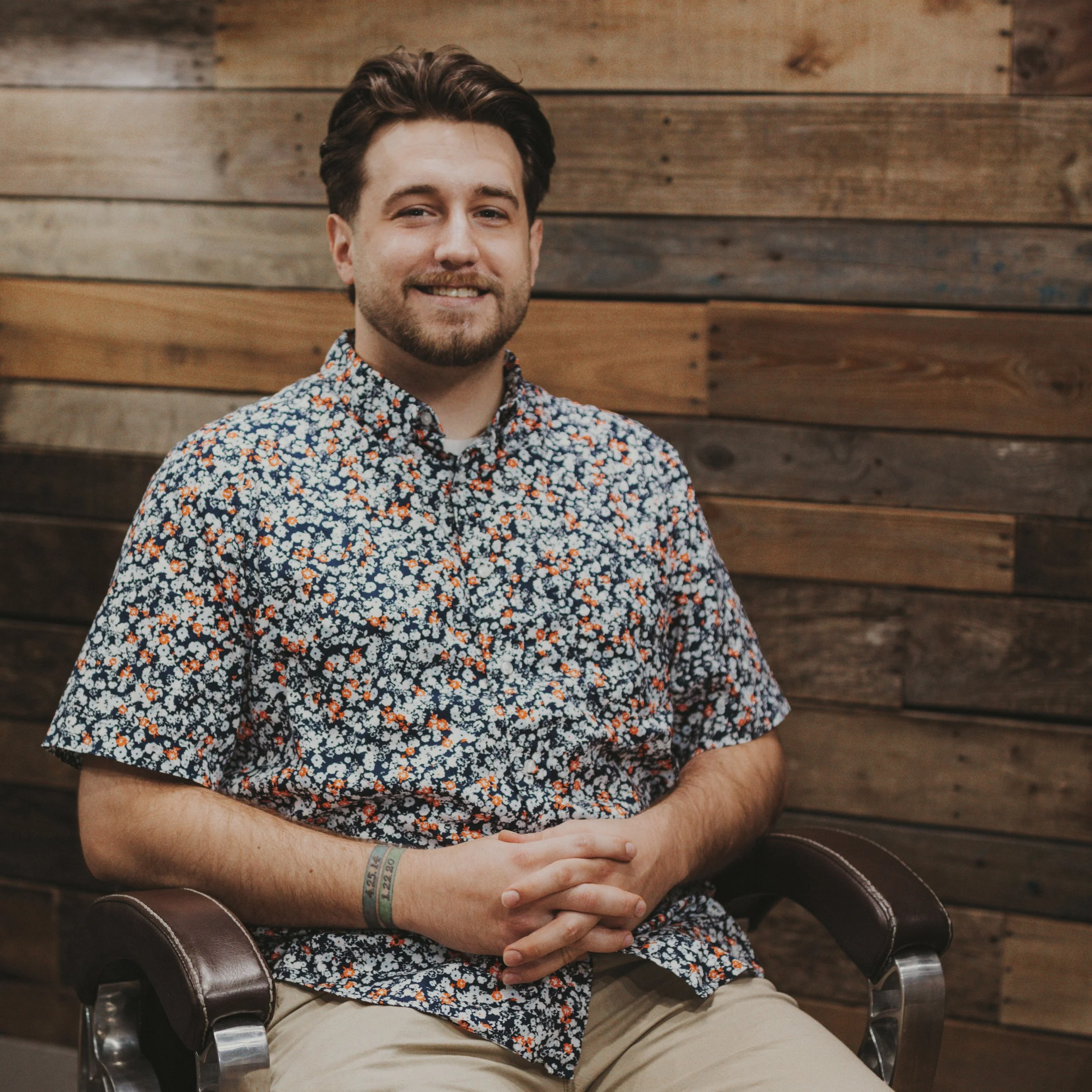 A man with dark brown hair, light skin, and facial hair sitting on a brown leather chair with armrests, wearing a floral-patterned shirt and khaki pants. He is smiling slightly with his hands clasped on his lap, and a hospital wristband is visible on his right wrist. The background consists of a wall made of horizontal wooden planks.