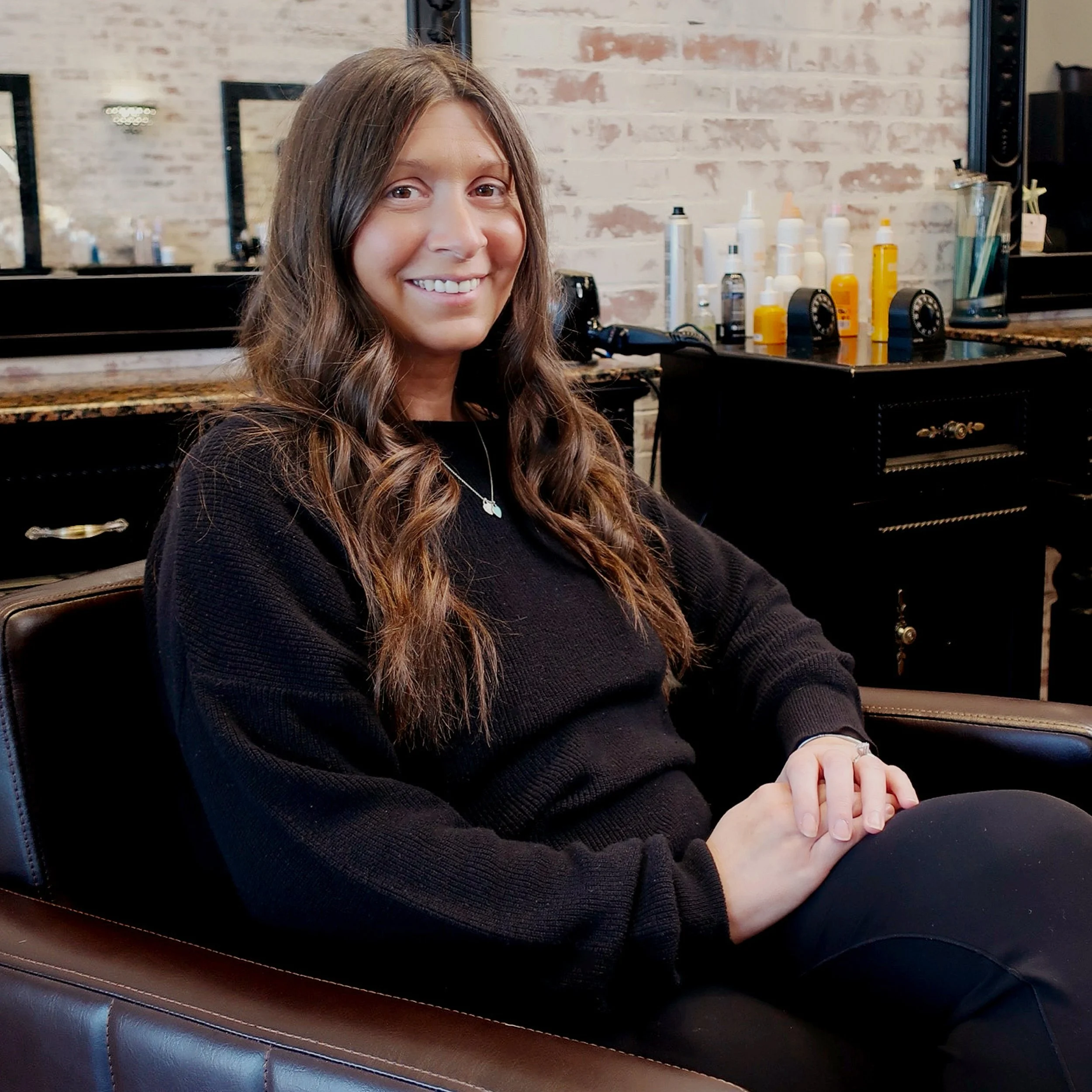 A woman with long, wavy brown hair, smiling and sitting in a salon with a brick wall and mirrors behind her. She is wearing a black sweater and a silver necklace.