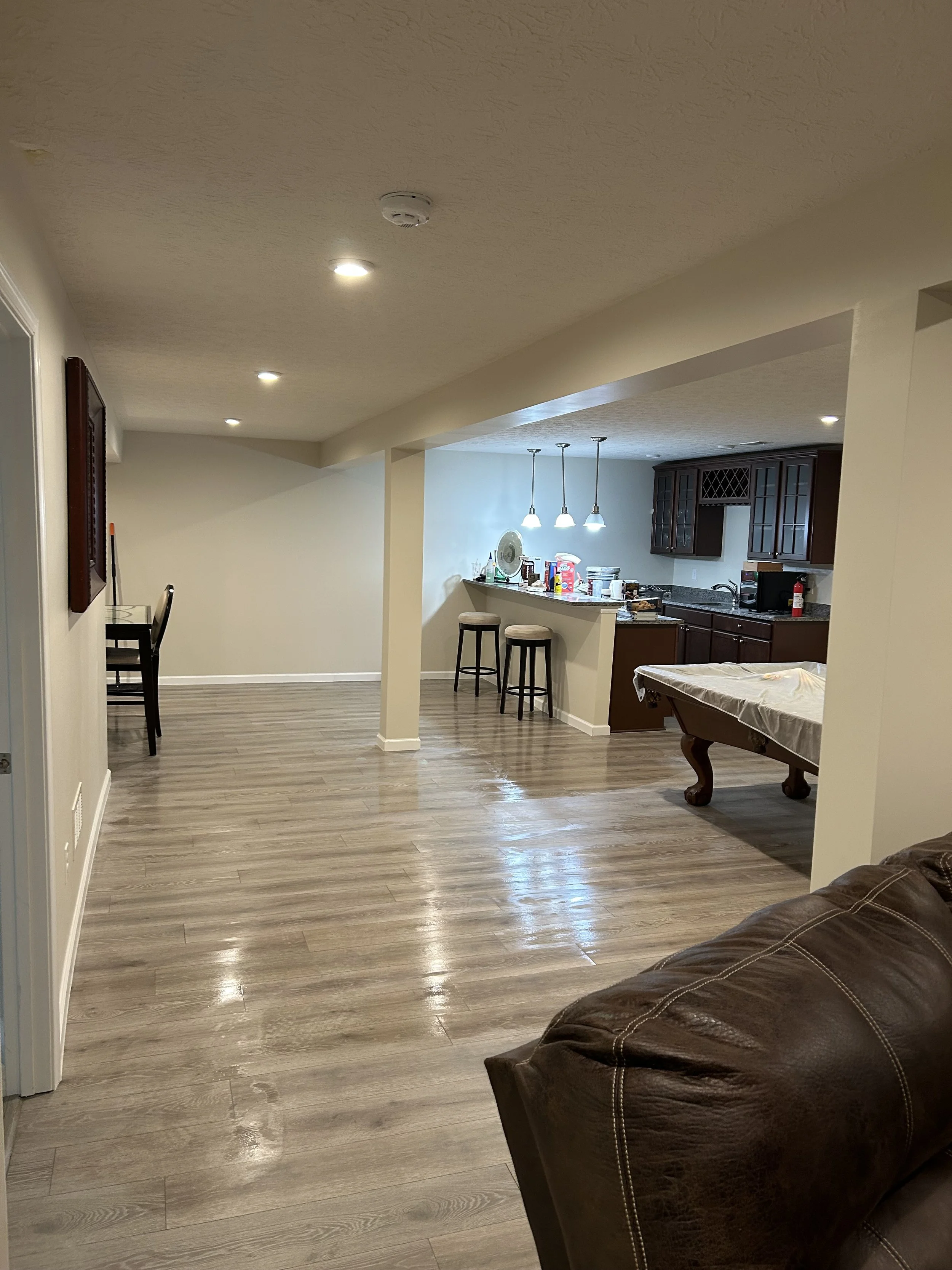 A living room with wood laminate floors, cream walls, a brown leather couch, a picnic table on the right, and a kitchen area with dark wood cabinets, a bar with two stools, pendant lights, and various items on the countertop.