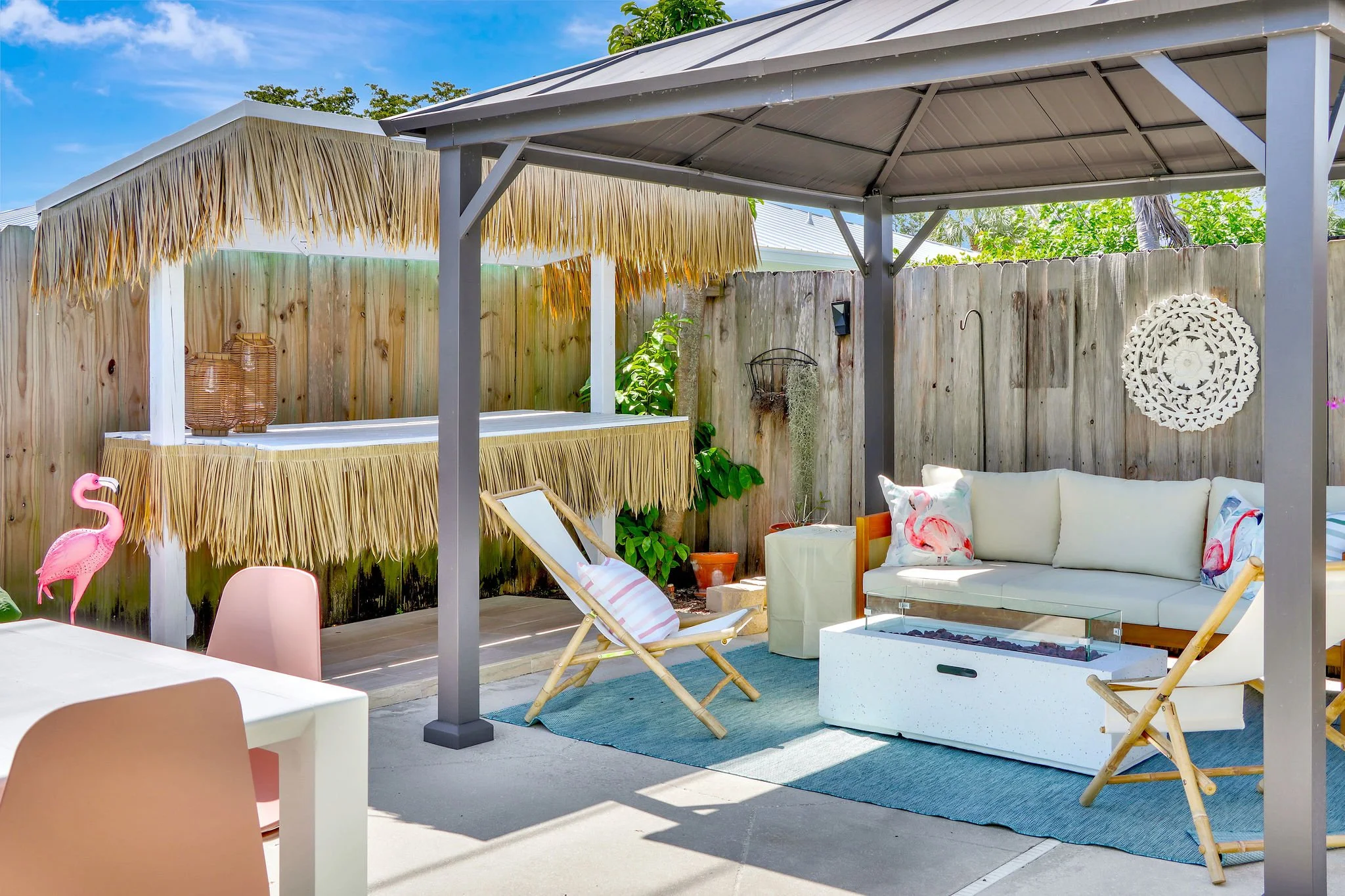 An outdoor patio area with a thatched roof canopy, a wooden fence, a white cushioned sofa, pink flamingo decorations, and various potted plants under a partly cloudy sky.