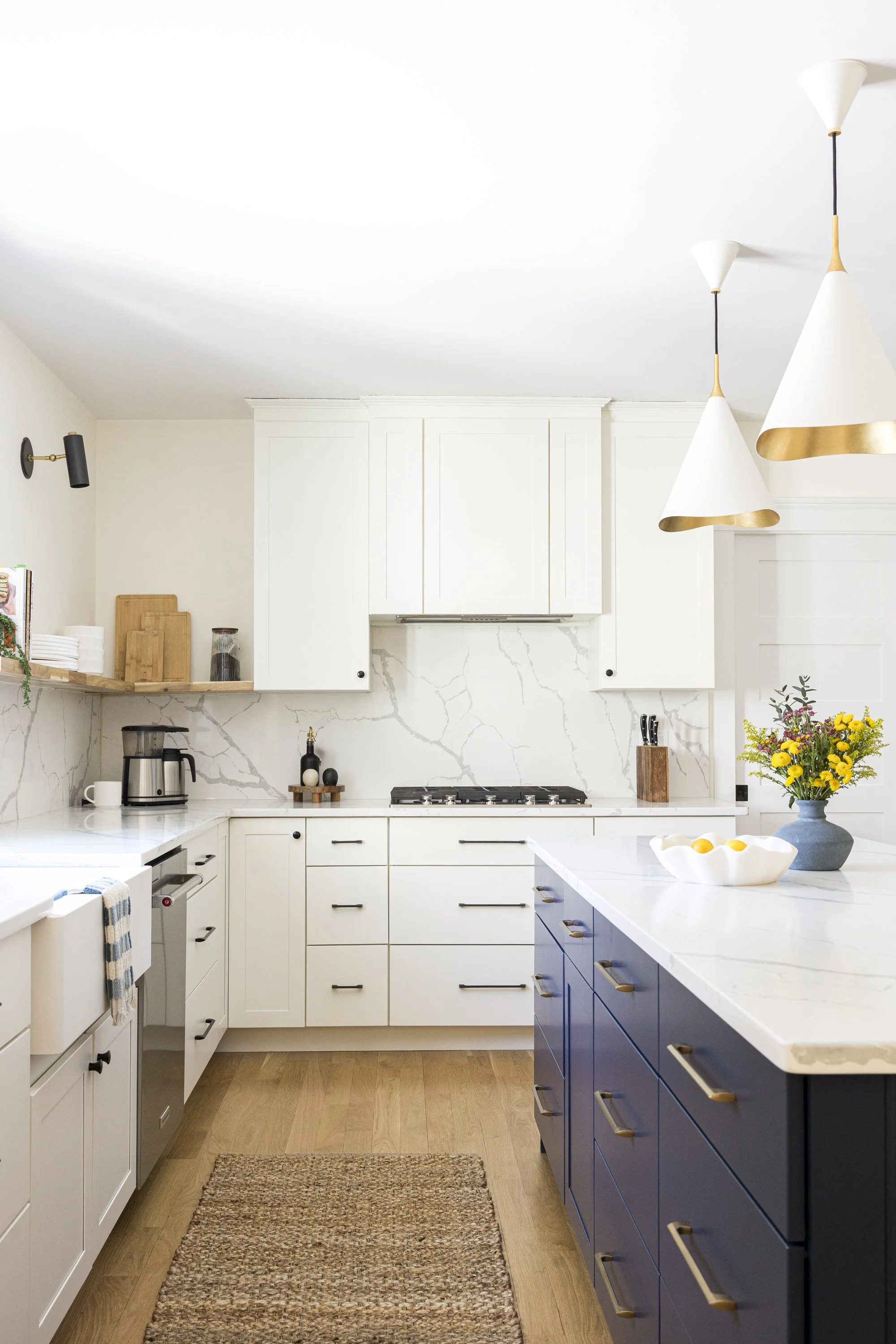 A modern kitchen with white upper cabinets, blue lower cabinets, marble countertops, pendant lights, a flower vase, and a kitchen rug.