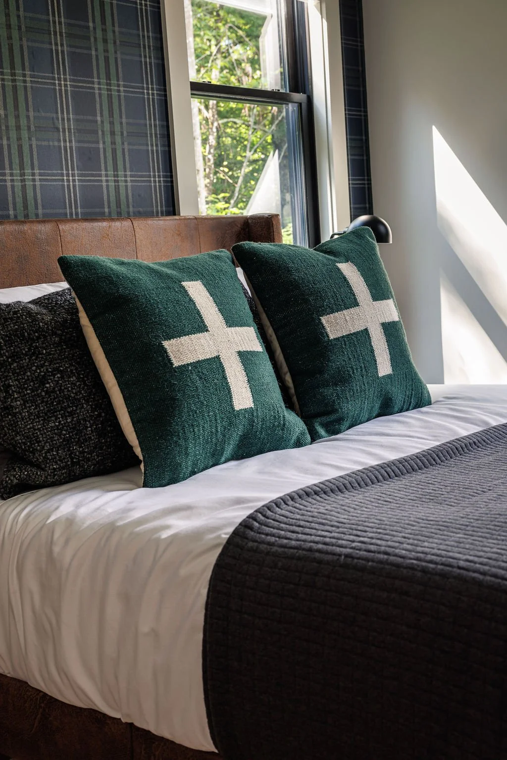 A cozy bedroom with a bed featuring white sheets, a dark gray quilted blanket, and two green pillows with white crosses. A window behind the bed lets in natural light, and a black bedside lamp is mounted beside the window.