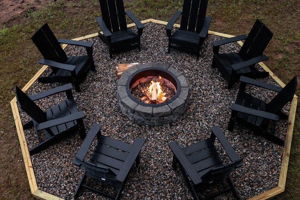 A circle of eight black Adirondack chairs surrounds a fire pit with a small fire, on gravel ground within a wooden border, outside on green grass.