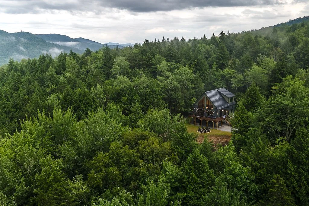 A house with a metal roof in a lush green forest with mountains and mist in the background.
